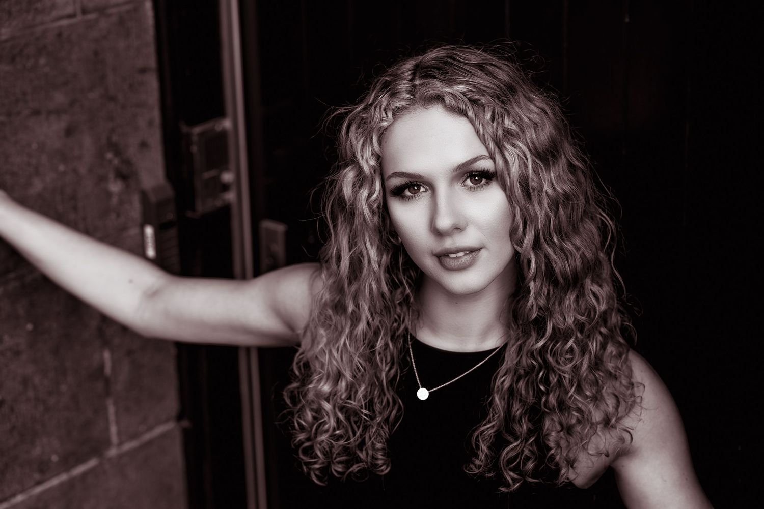 Black and white portrait of a young woman with curly hair leaning against a door in dramatic lighting.