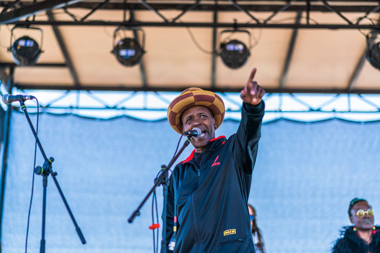 An energetic performer in a hat points skyward while singing at an outdoor Jamaican music festival stage.