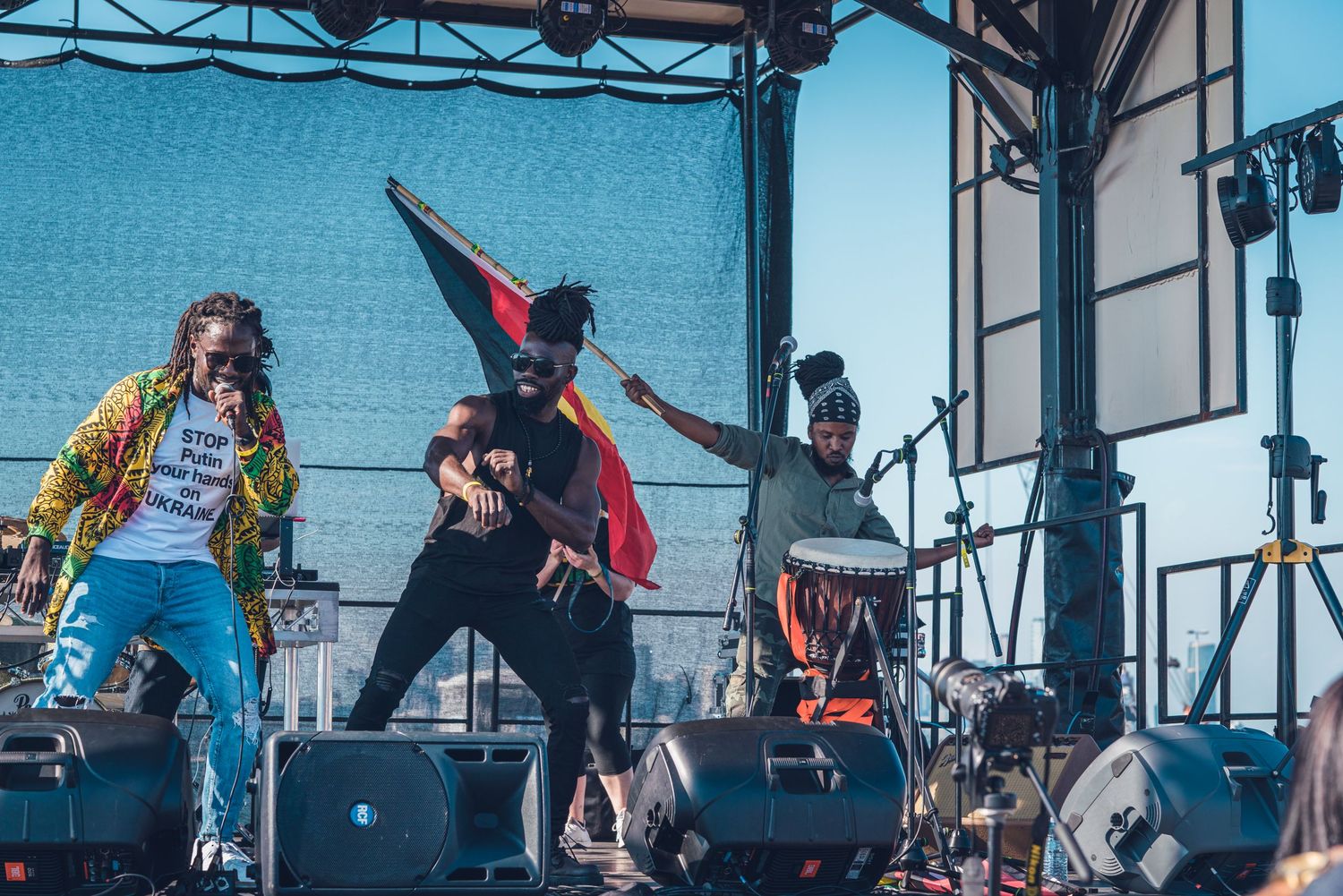 Energetic performers on outdoor festival stage with colorful outfits and city skyline backdrop.