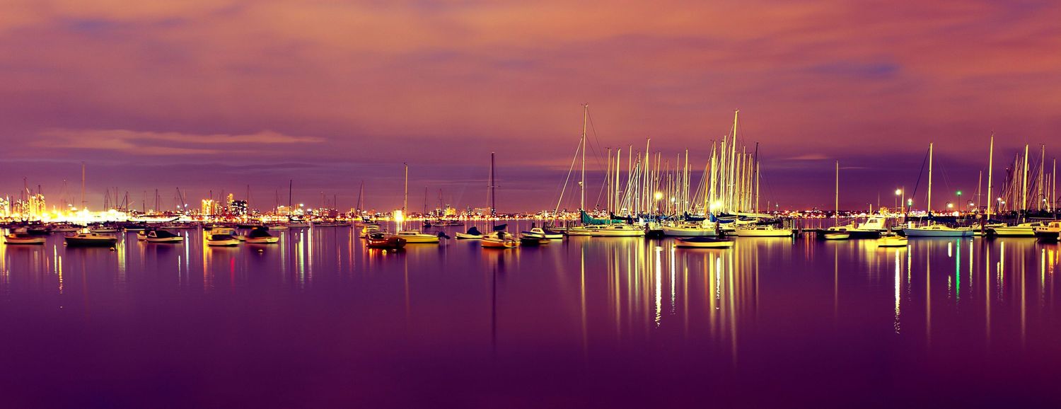A breathtaking purple dusk panorama of a marina with sailboats and city lights reflecting on still water.