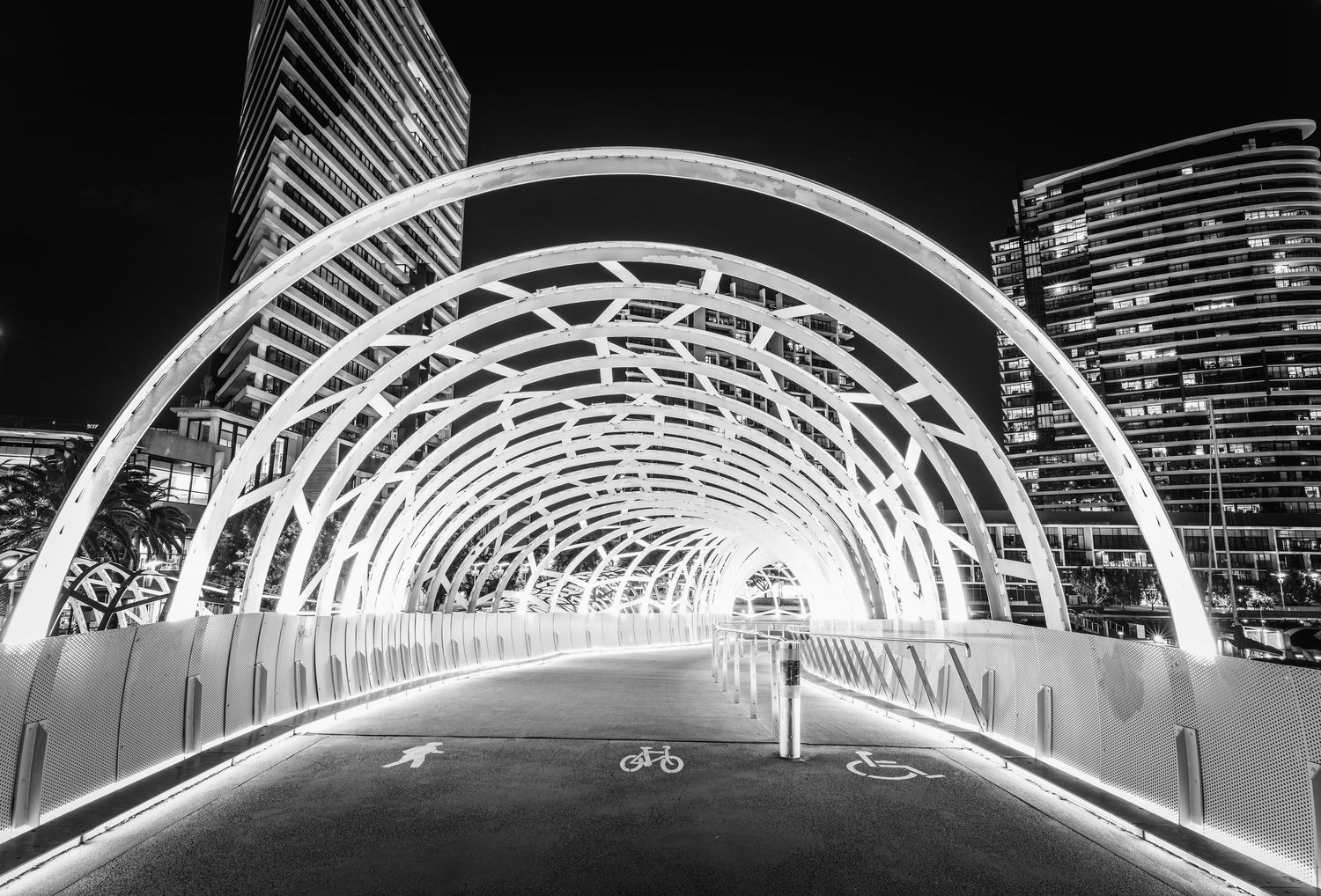 Black and white night photo of illuminated arched pedestrian bridge in urban cityscape.