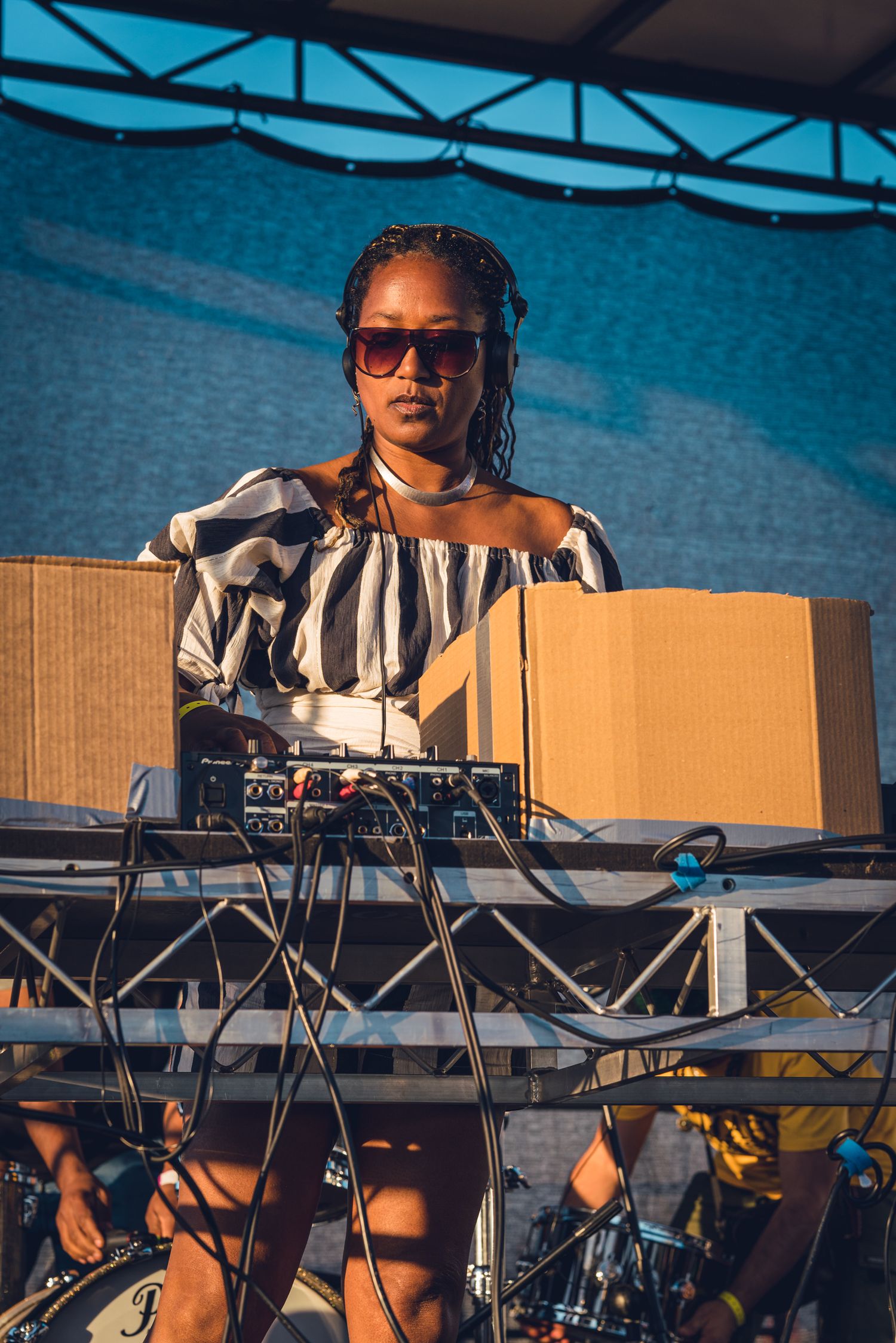 Female DJ in striped off-shoulder top performing behind equipment at outdoor festival.