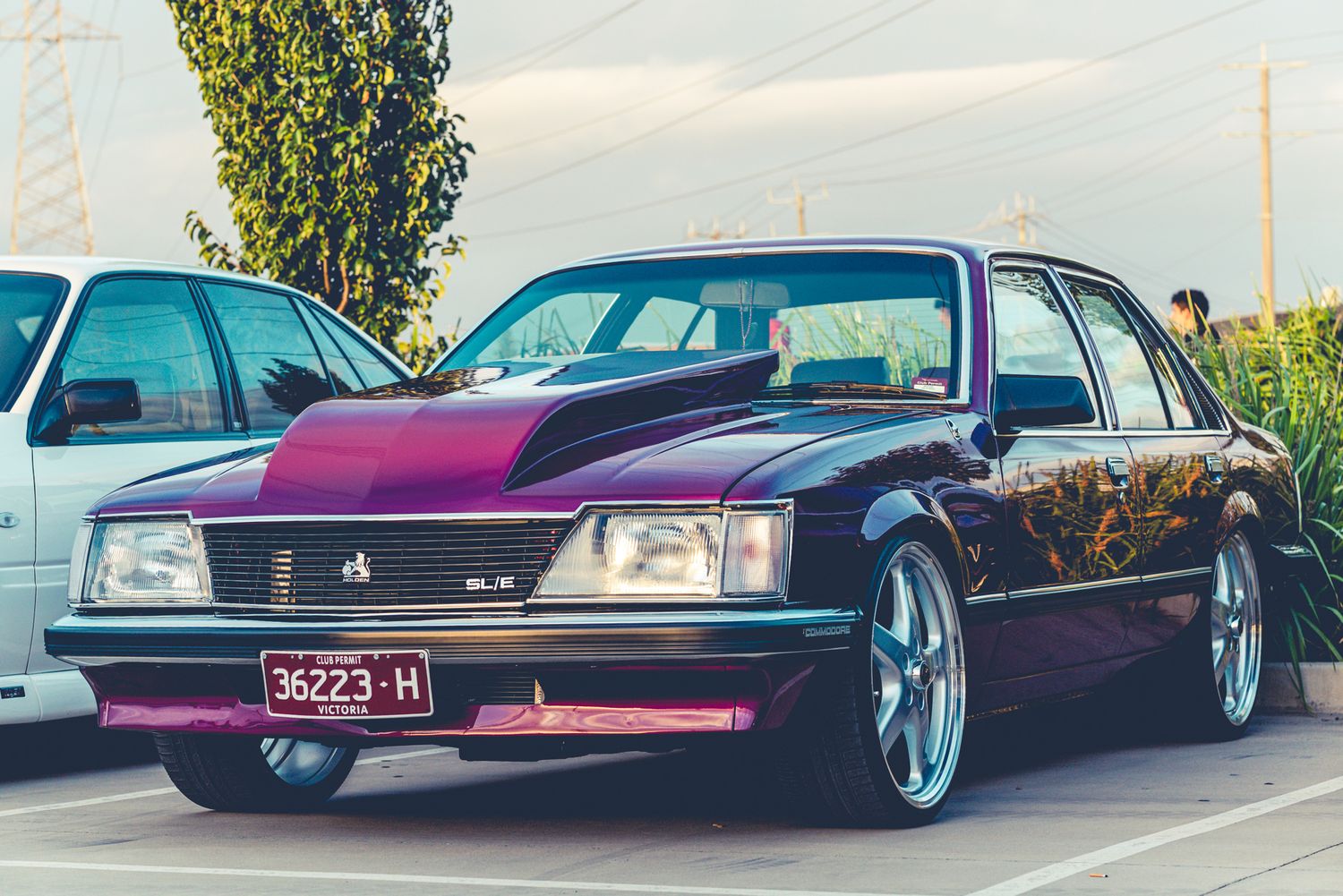 Side view of a lowered purple custom Holden SL/E with pink hood accent and chrome wheels at a car show.
