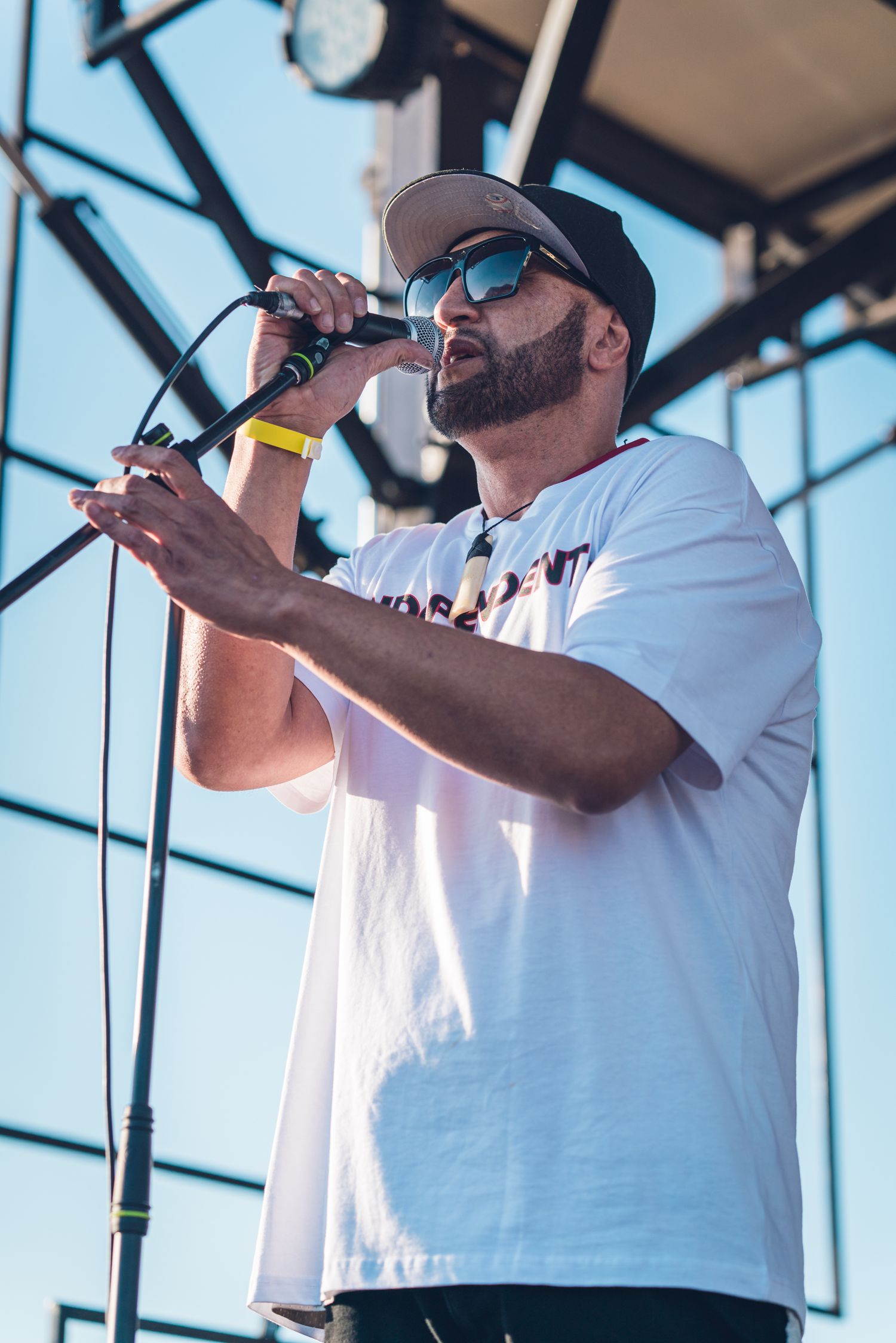 Bearded male rapper in white t-shirt and snapback cap performing into microphone on outdoor stage.