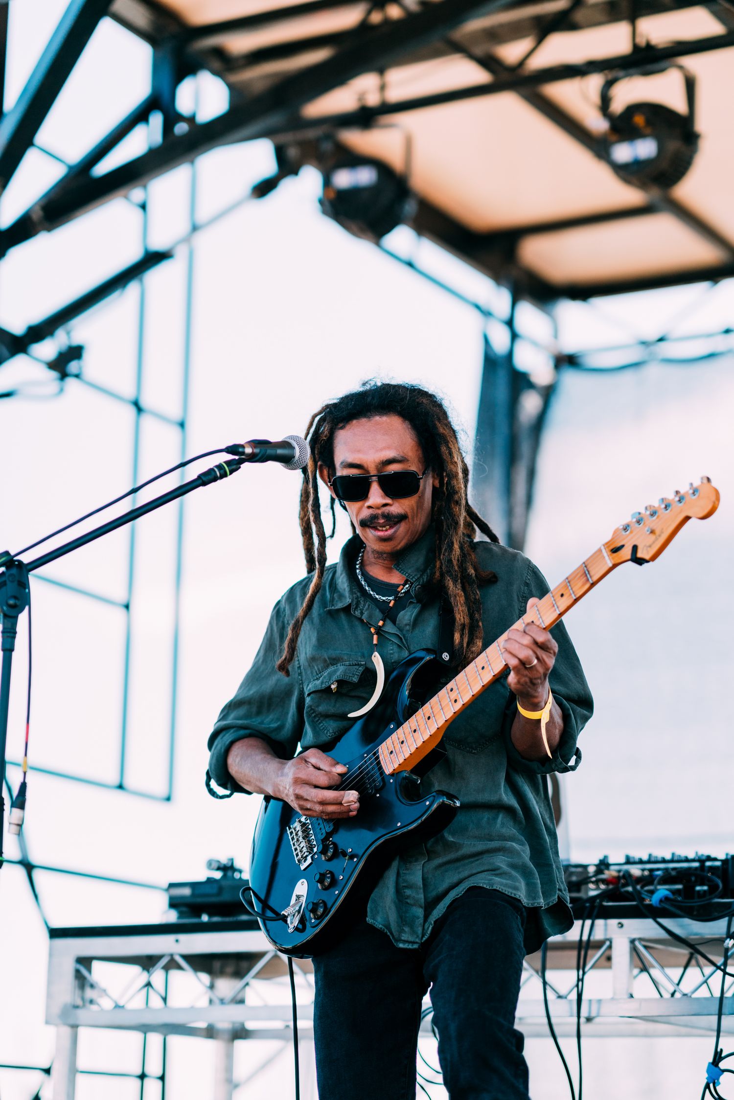 A dreadlocked musician in sunglasses plays a blue electric guitar on an outdoor festival stage.