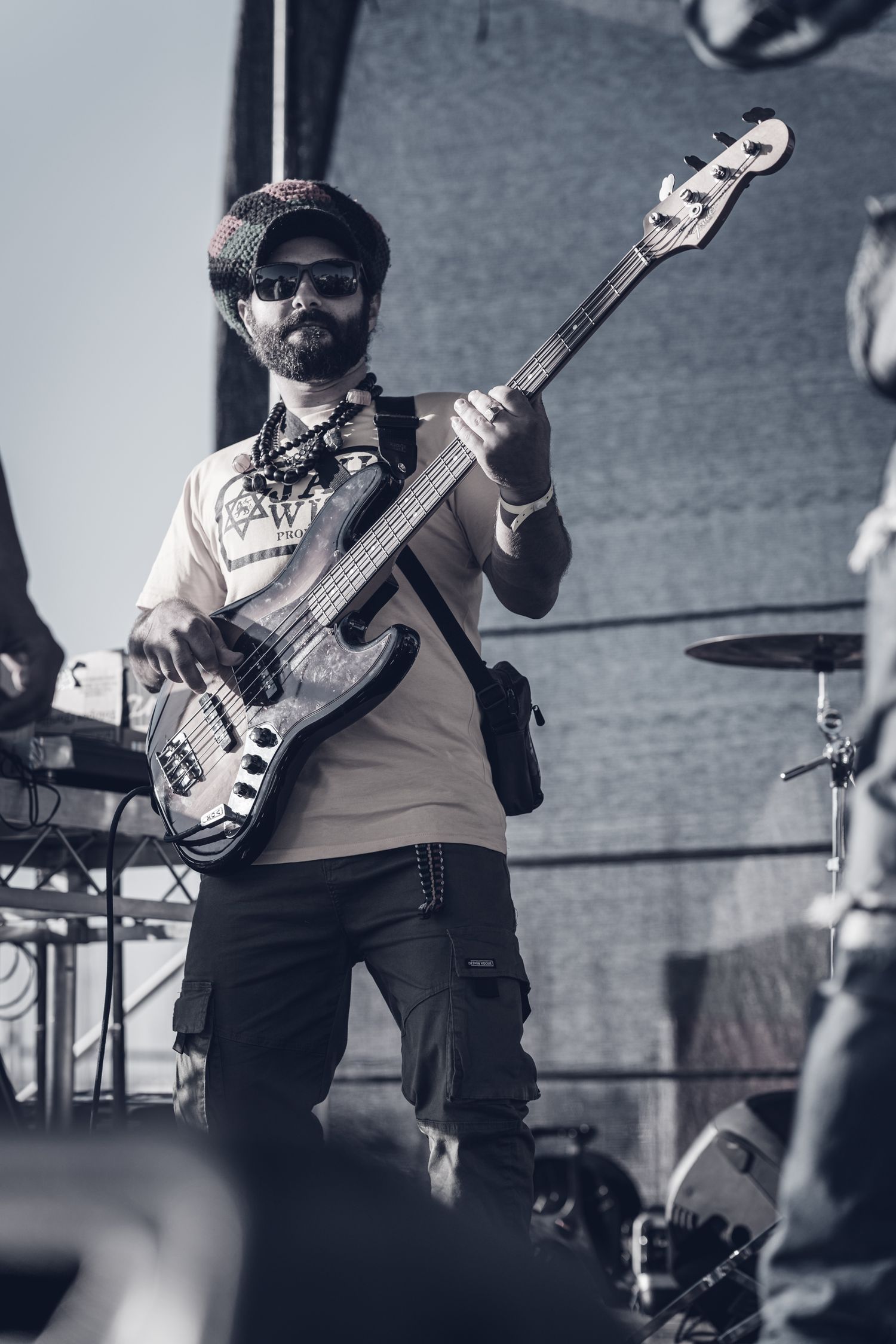 Male bassist with beard playing electric bass guitar on outdoor festival stage in desaturated tones.