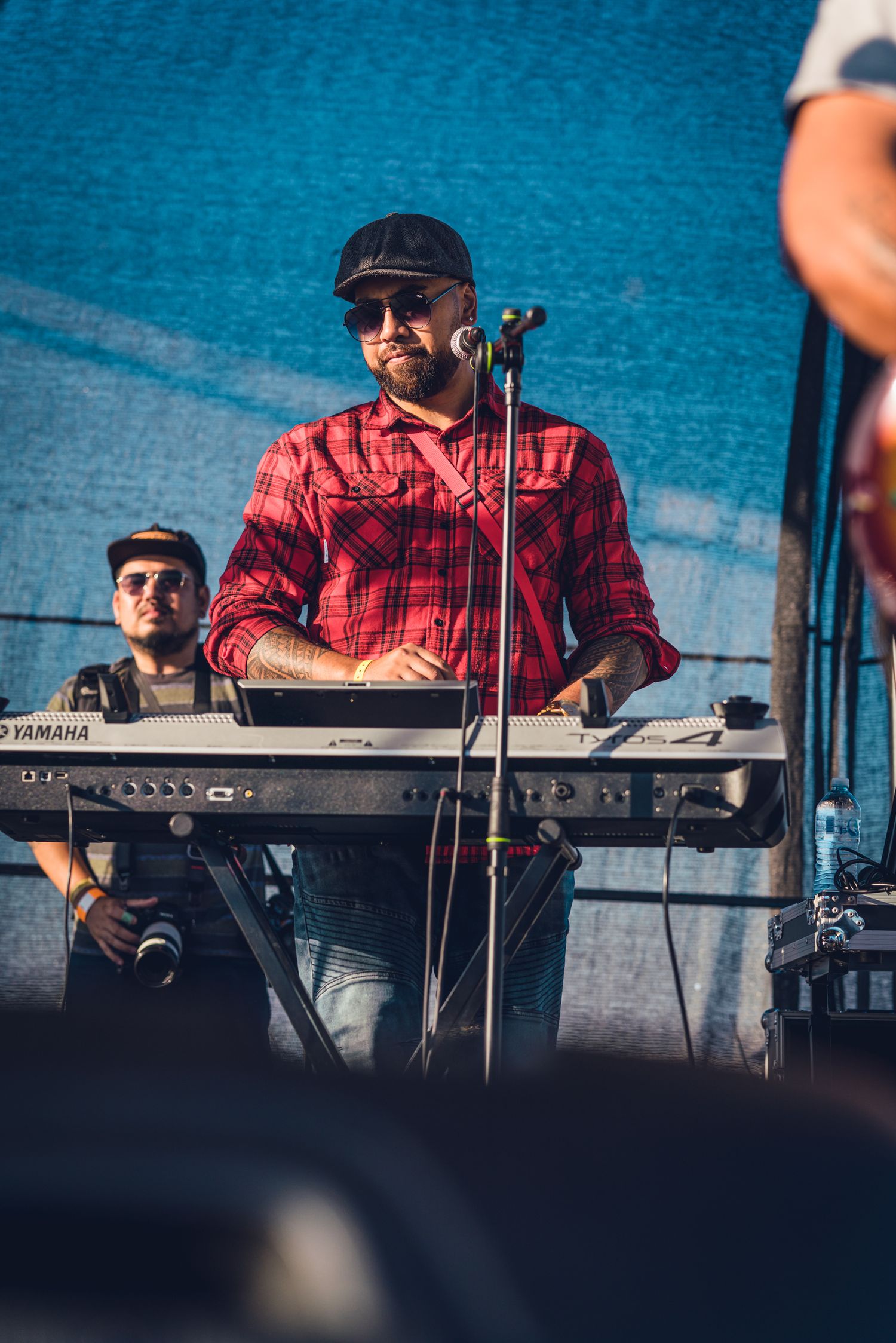 Smiling male keyboardist in red flannel shirt and cap performing on Yamaha keyboard at outdoor festival.
