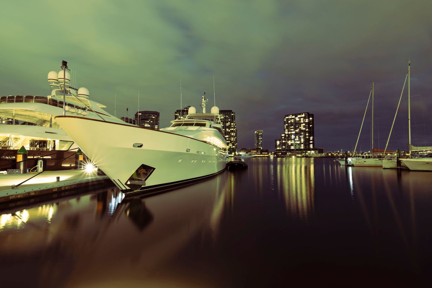 Luxury white superyacht moored at a marina at night with glowing city skyline in the background.