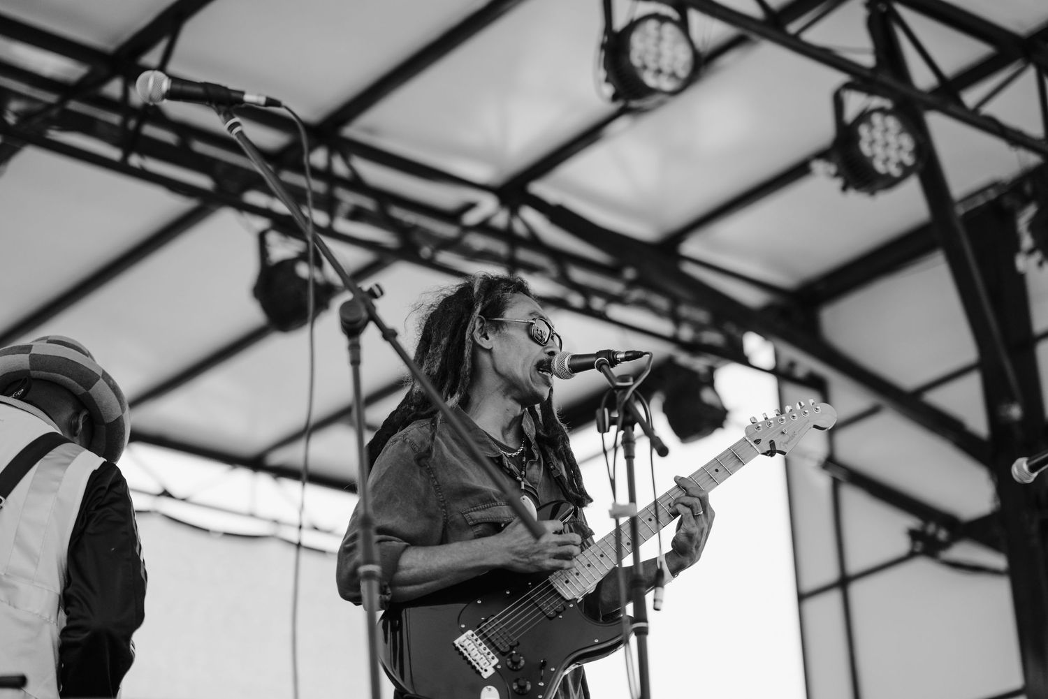 Black and white photo of a dreadlocked guitarist singing into a microphone on an outdoor festival stage.