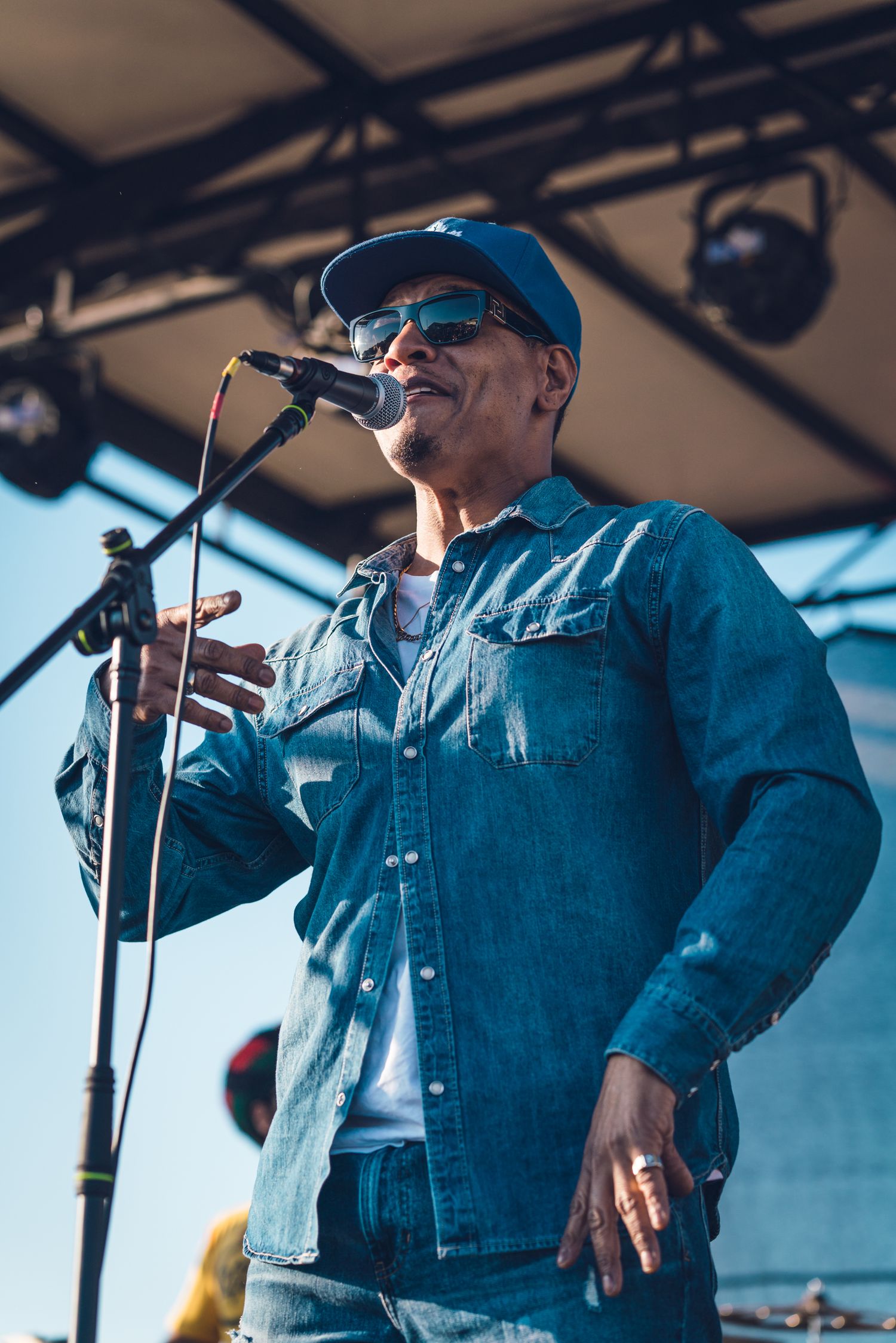 Male vocalist in denim jacket and blue cap singing into microphone on outdoor festival stage.