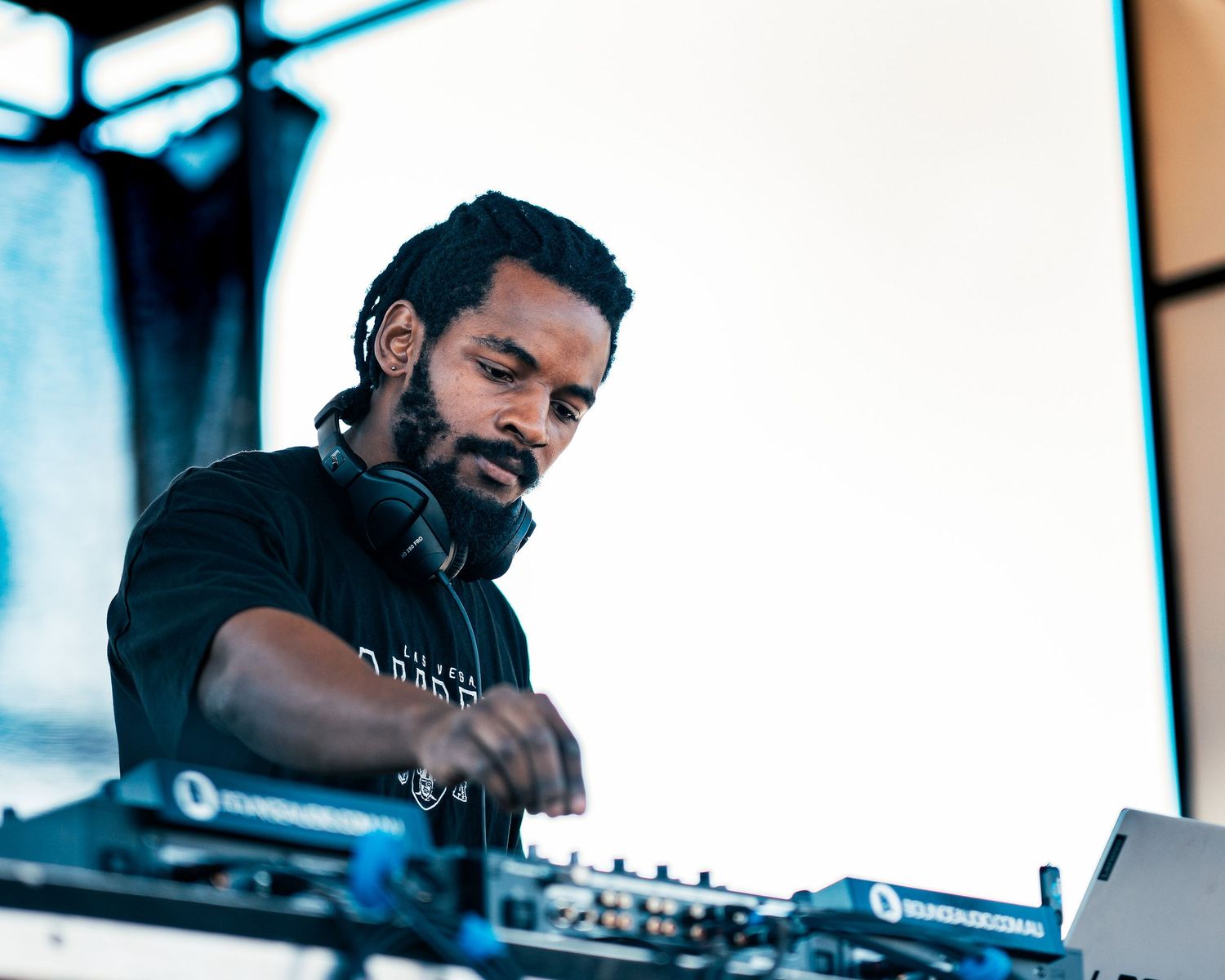 A focused male DJ with headphones around his neck works the turntables at an outdoor music festival.