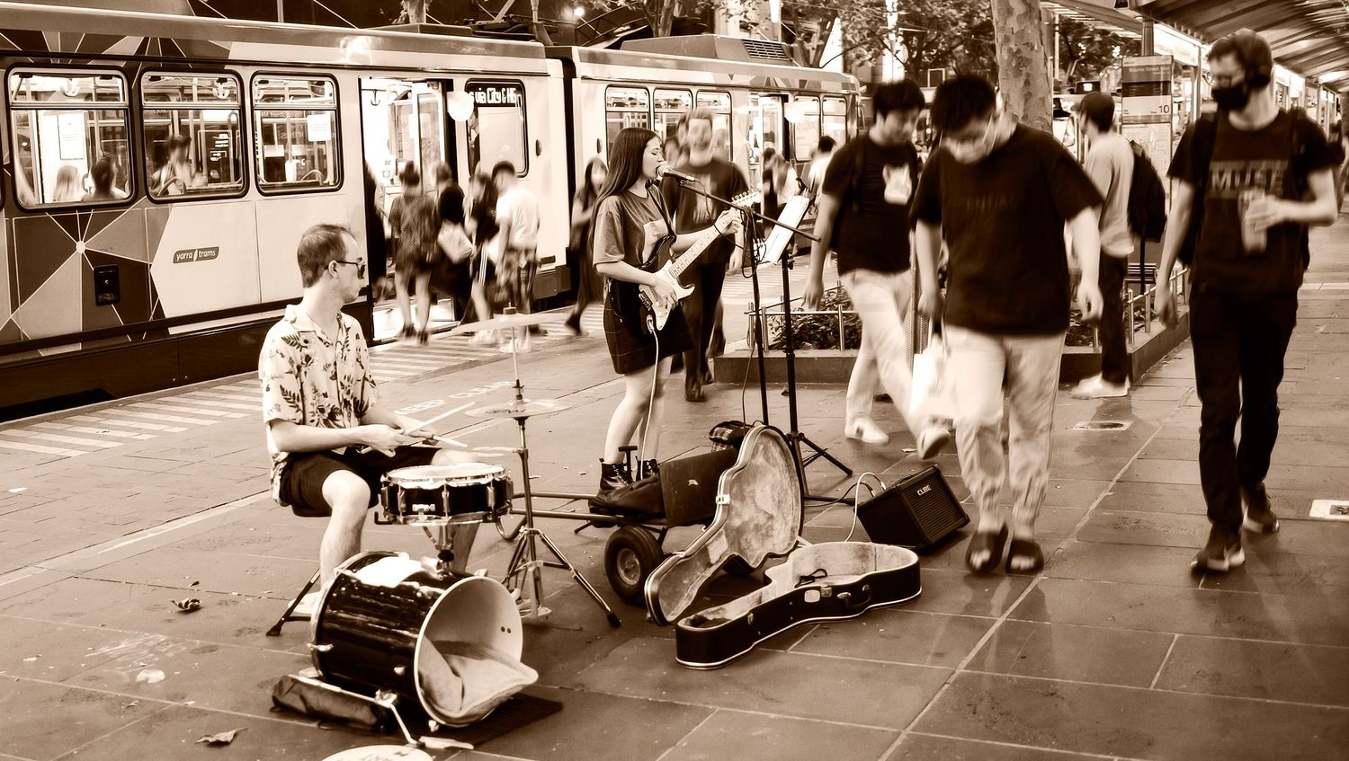 Sepia-toned street band playing drums and guitar on a bustling city tram station sidewalk.