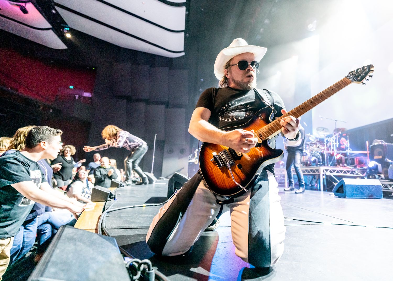 A guitarist kneels on stage in a white cowboy hat, playing an electric guitar at a live concert.