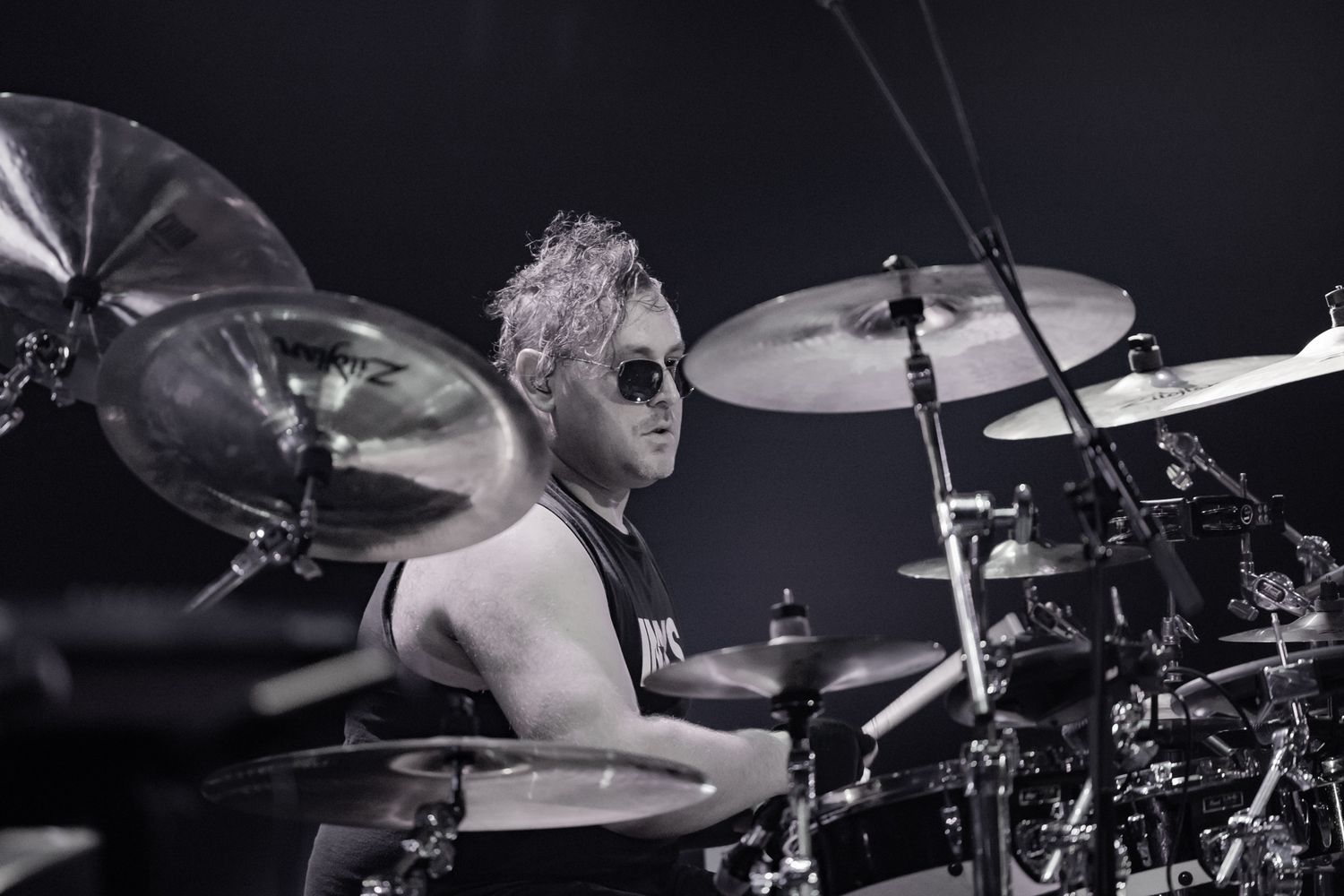 Black and white close-up of a curly-haired drummer wearing sunglasses performing energetically on a drum kit.