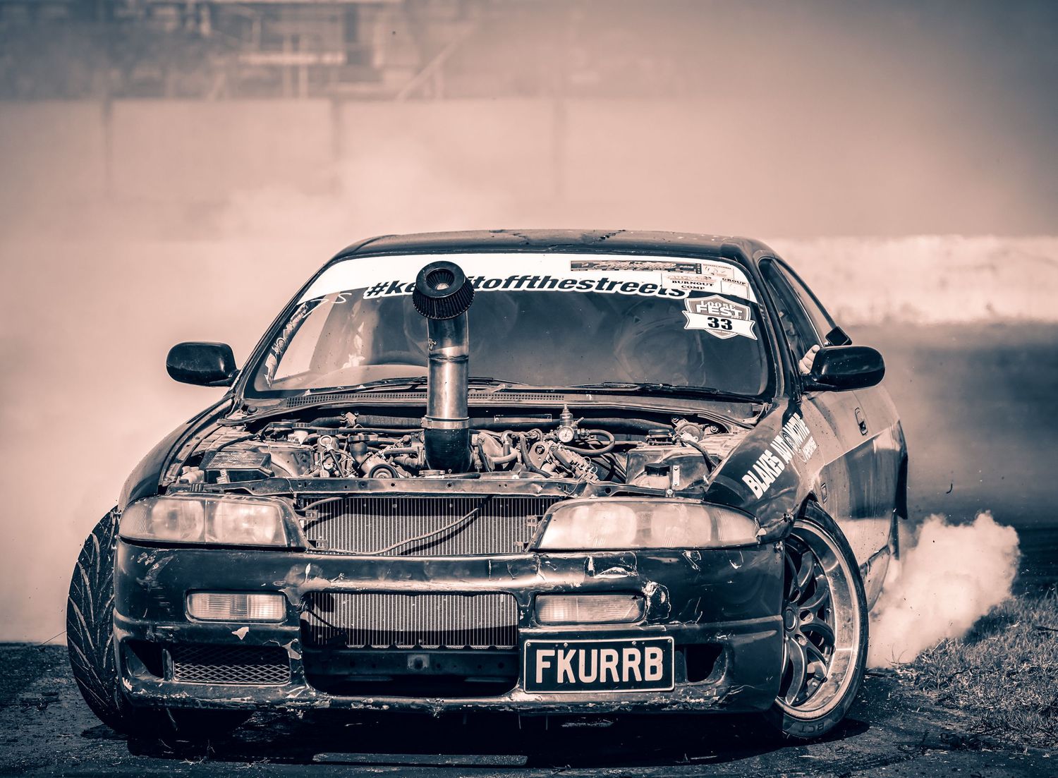 A modified drift car with a blower engine performs a smoky burnout on a track.