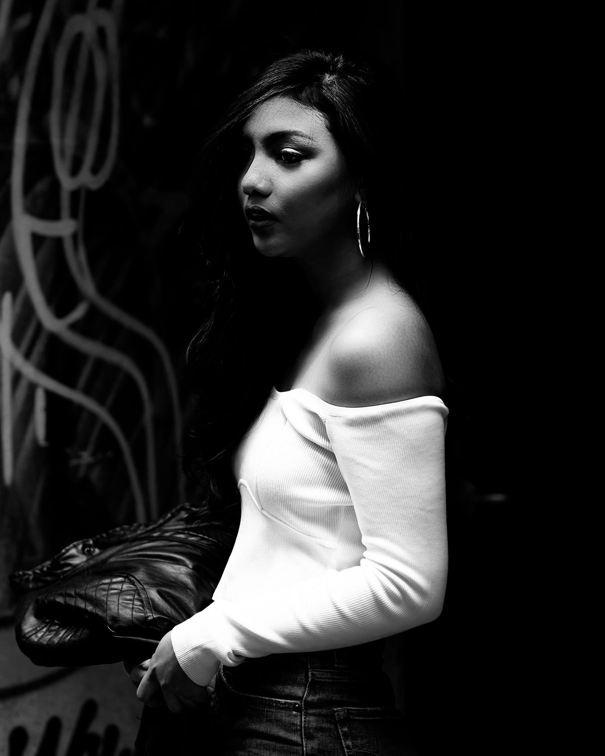 Black and white portrait of a woman in off-shoulder white top holding a leather bag against a graffiti wall.