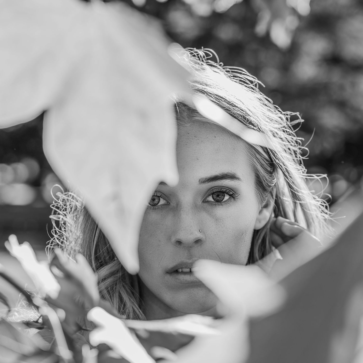 Black and white portrait of blonde woman peering through large tropical leaves outdoors.