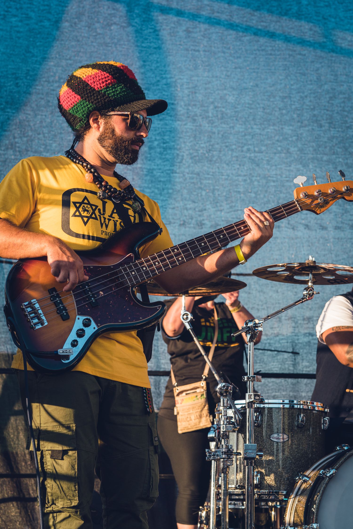 Bassist in Rastafarian hat and yellow shirt playing bass guitar at outdoor festival.