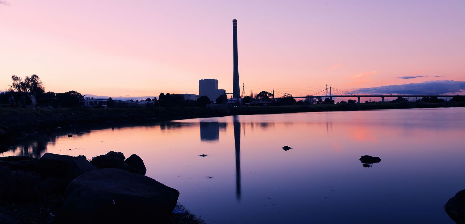 Industrial chimney stack reflecting in calm river at pink and purple sunset dusk, panoramic waterfront view.
