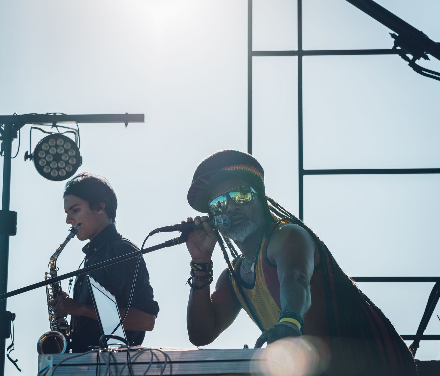 Two musicians performing on sunny outdoor stage, one playing saxophone and one singing into microphone.