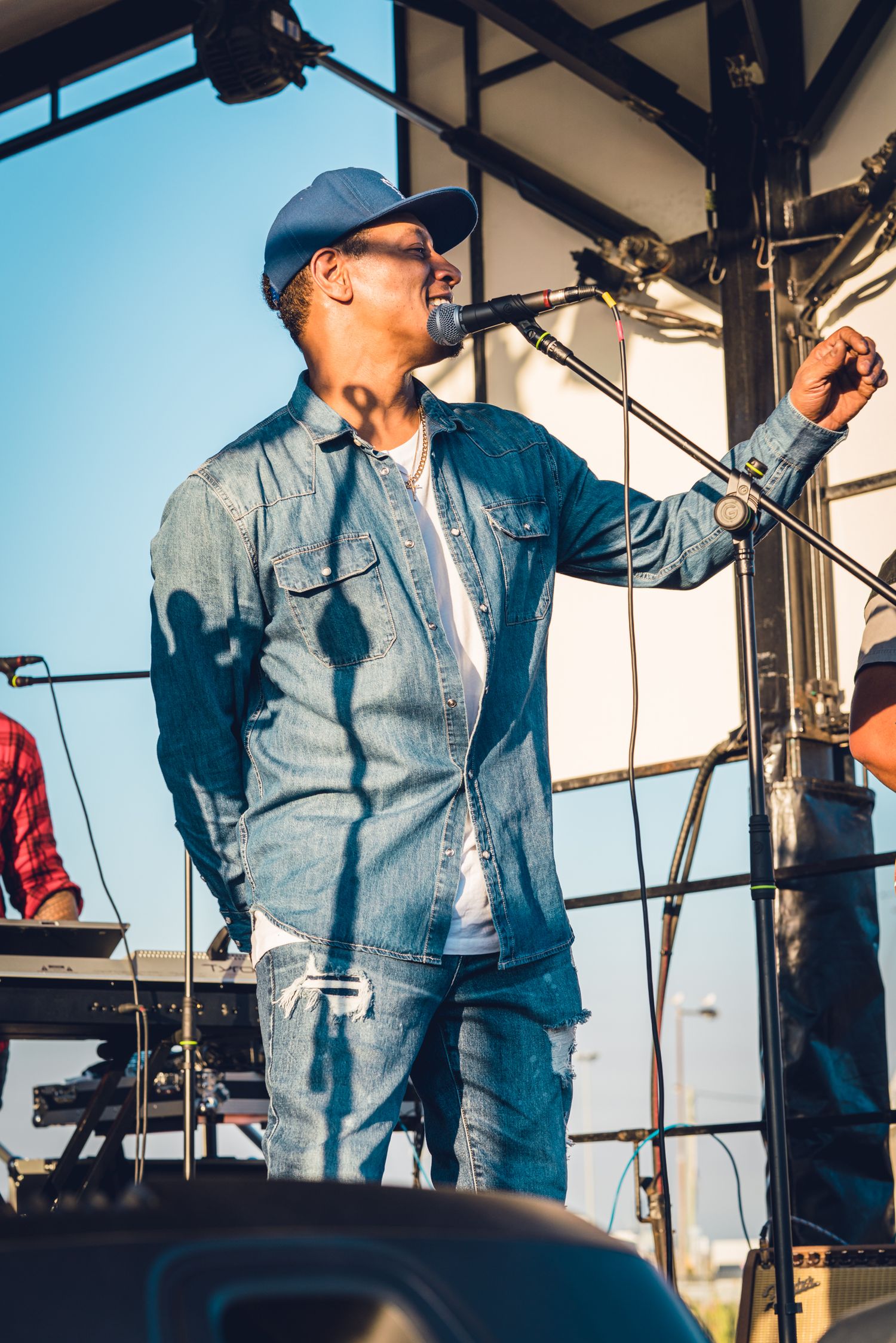 Singer in denim outfit performing on outdoor stage holding microphone under blue sky.