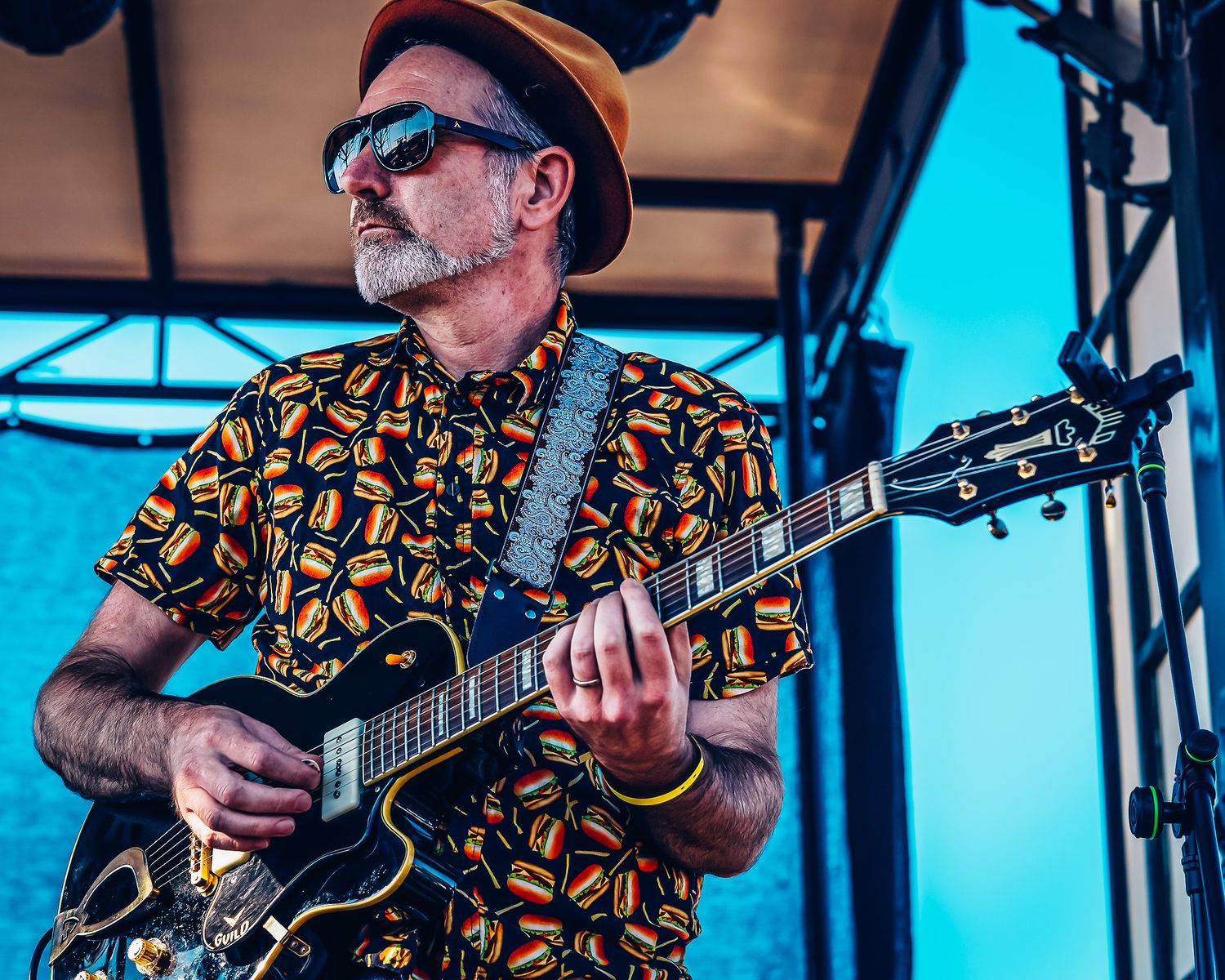 A guitarist in a colorful patterned shirt and hat performs on an outdoor stage at a music festival.