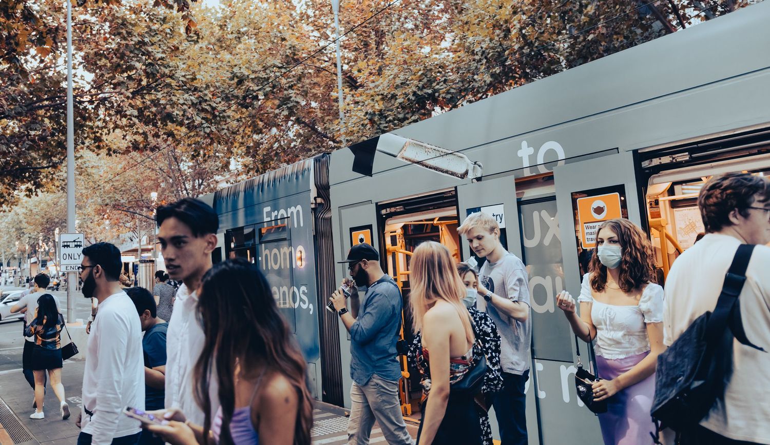 Busy urban street scene with crowds of people boarding a tram on a sunny tree-lined boulevard.