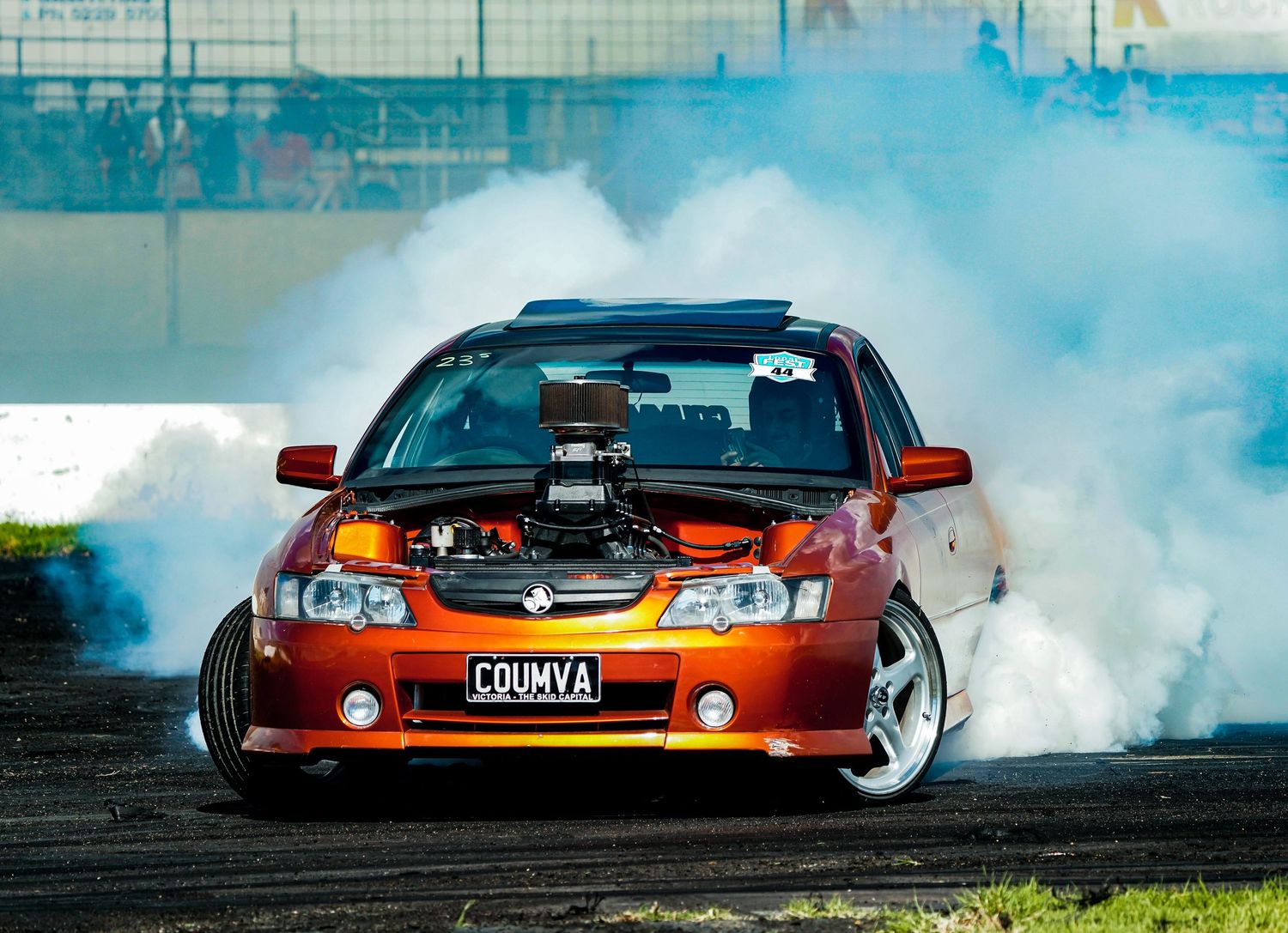 An orange Holden modified car performs a burnout surrounded by thick white smoke.