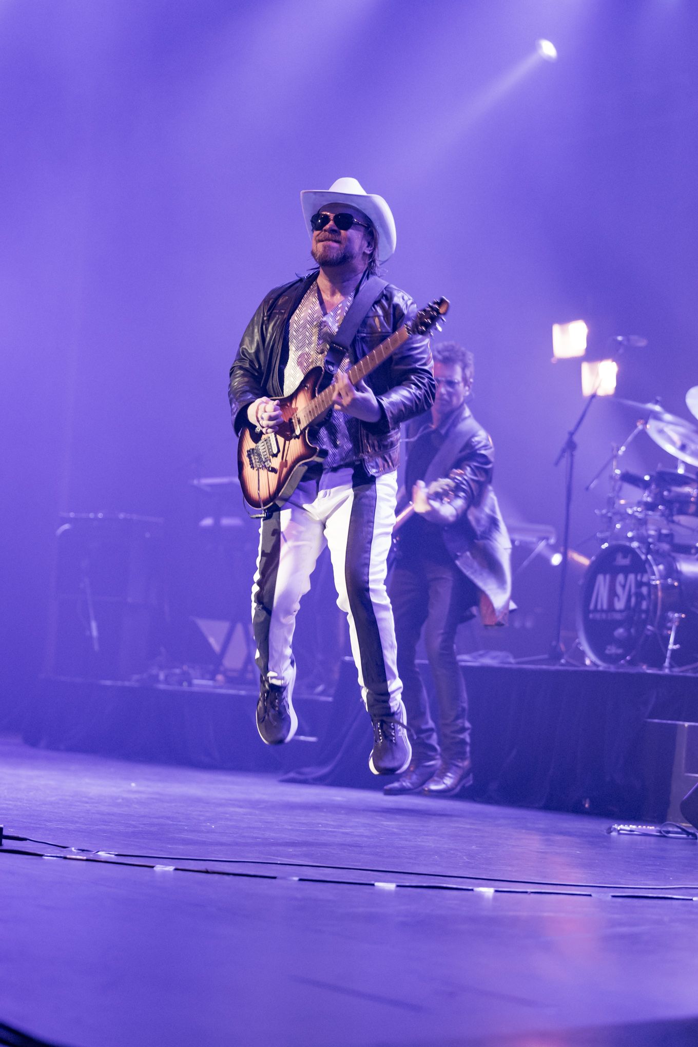 Energetic guitarist in a white cowboy hat jumping mid-air on a purple-lit concert stage.
