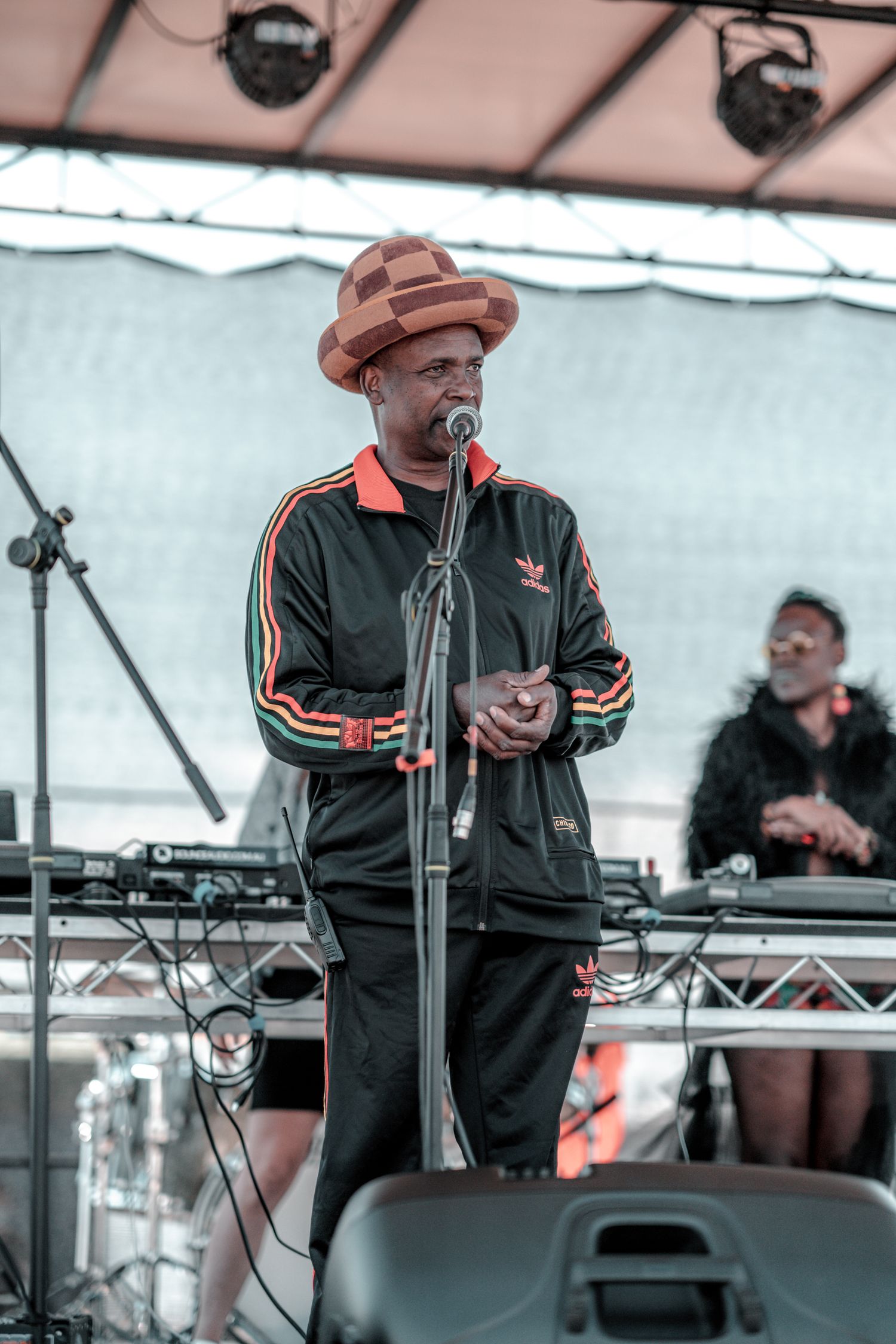 A man in a Adidas tracksuit and brown hat performs on stage at an outdoor Jamaican music festival.