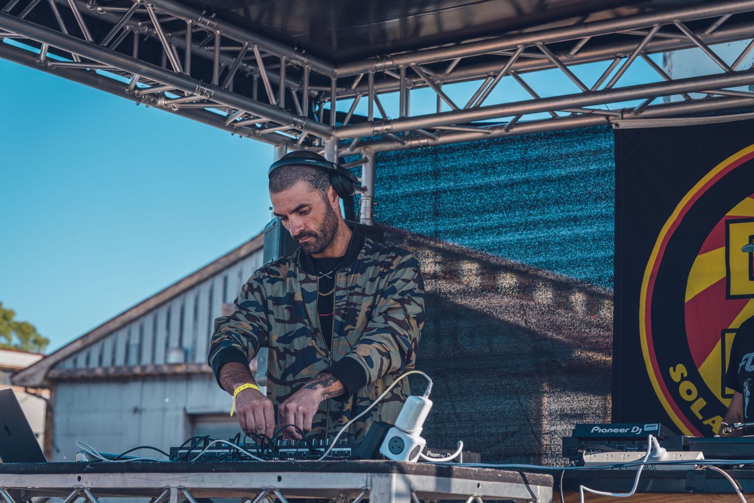 Male DJ in camouflage jacket mixing music at outdoor festival turntable setup.