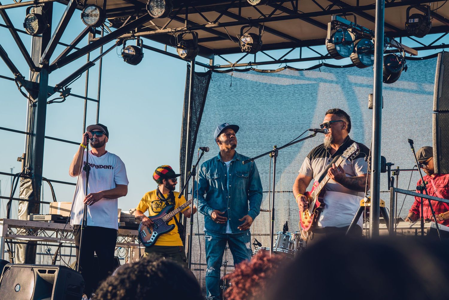 Four-piece band performing on outdoor festival stage under clear blue sky.