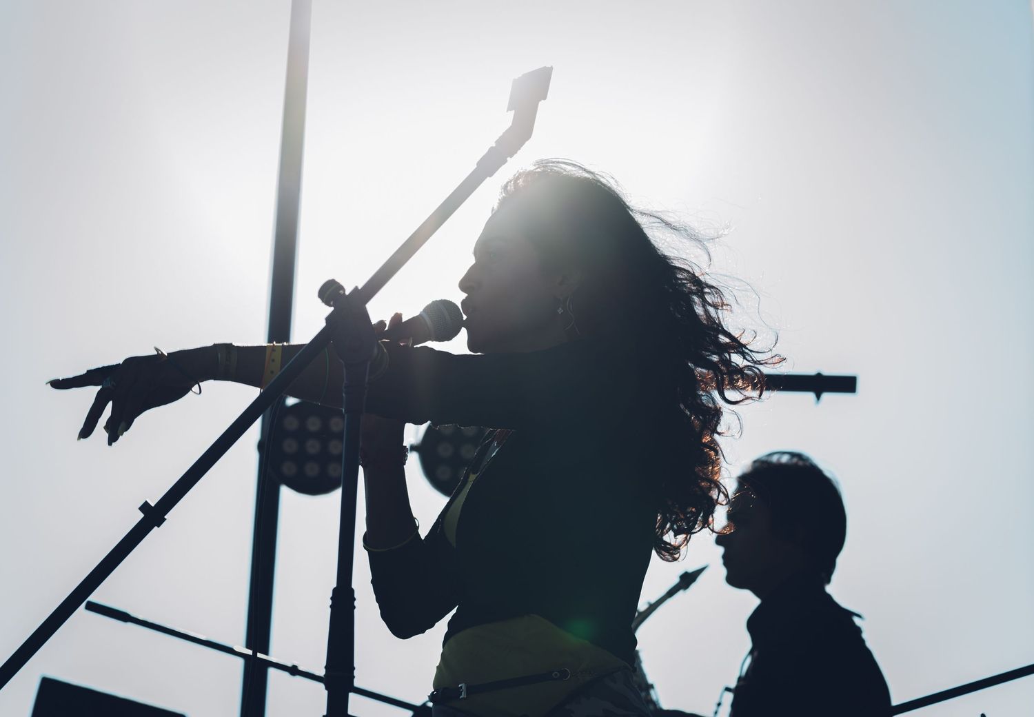 Silhouette of female vocalist singing into microphone with guitar neck visible against bright sun.