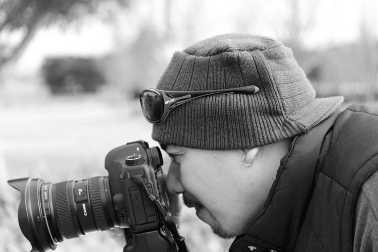 Black and white portrait of a photographer wearing a beanie and sunglasses, shooting with a DSLR camera outdoors.