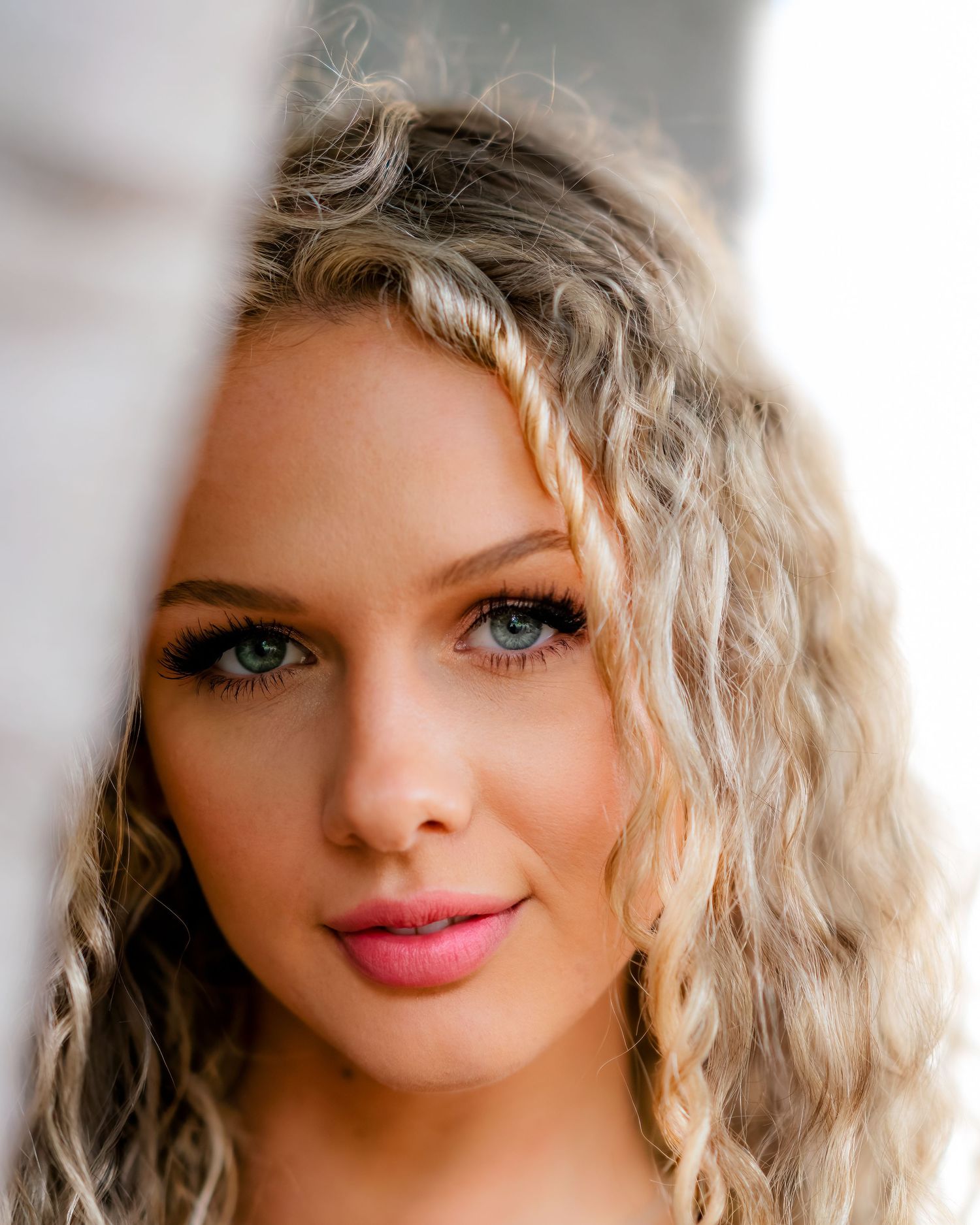 Close-up portrait of a blue-eyed blonde woman with curly hair and pink lips in natural lighting.