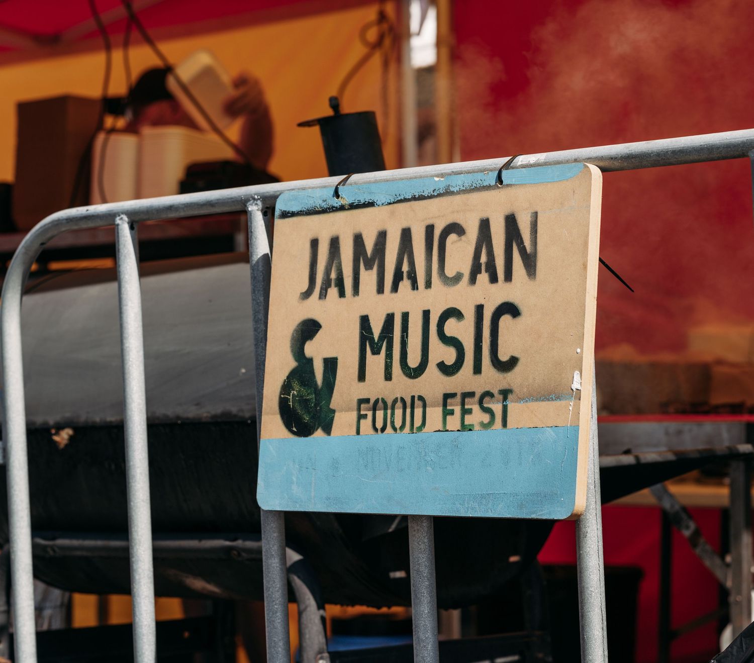 A vintage sign for the Jamaican Music and Food Fest displayed on a metal barricade at an outdoor festival.