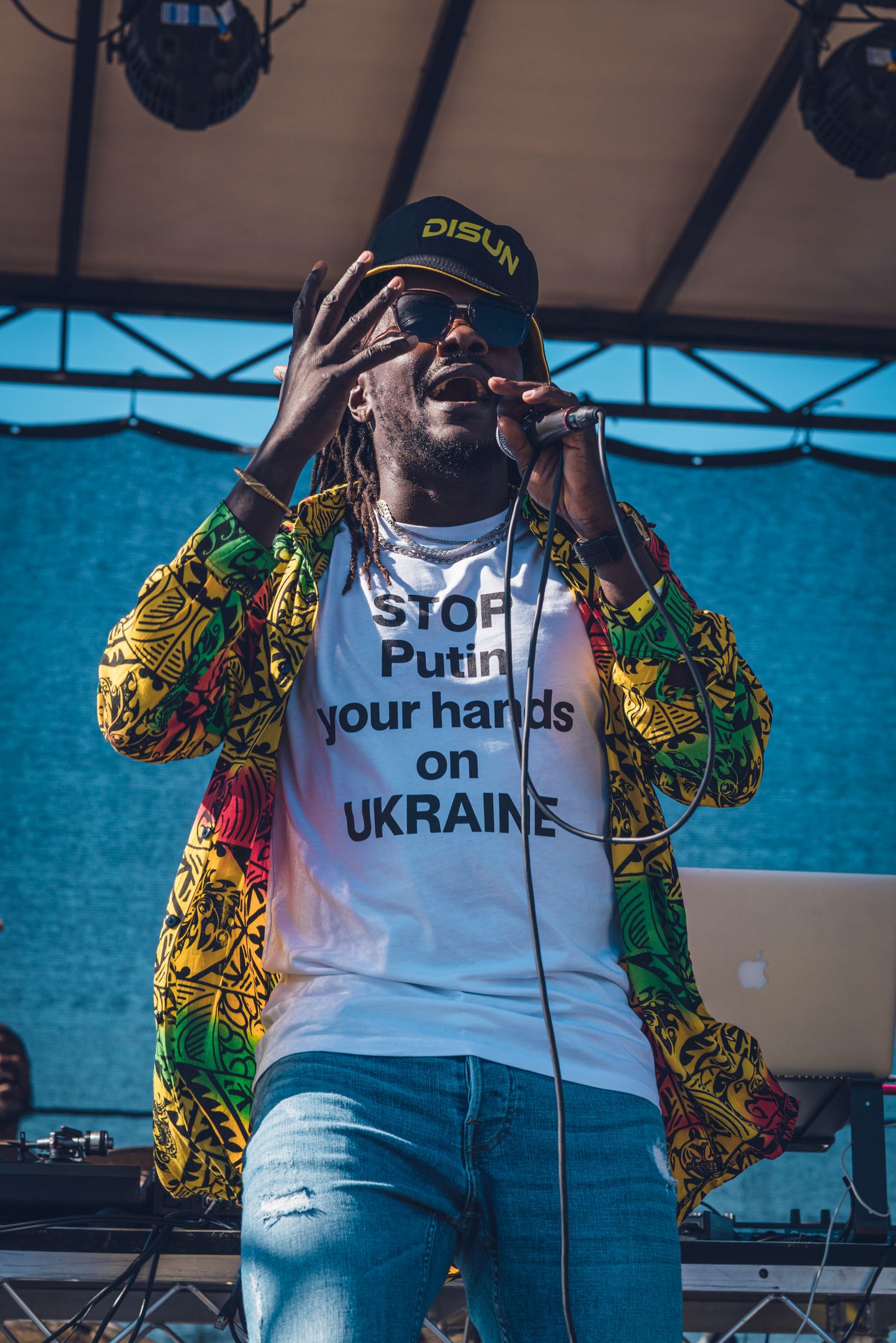 A reggae artist in a colorful jacket wears a Stop Putin Your Hands on Ukraine shirt while performing.