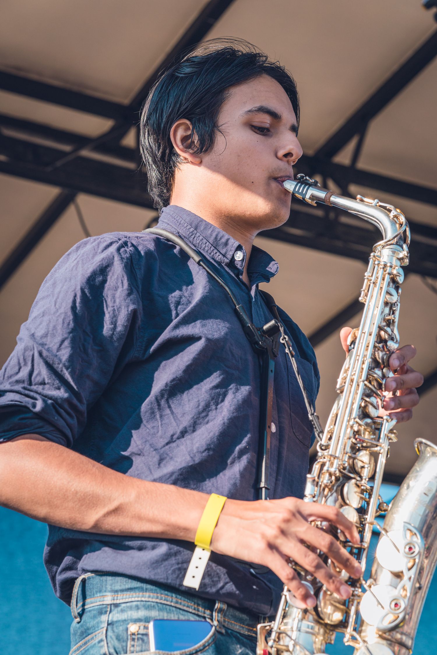 Young male saxophonist in blue shirt playing alto saxophone on outdoor festival stage.