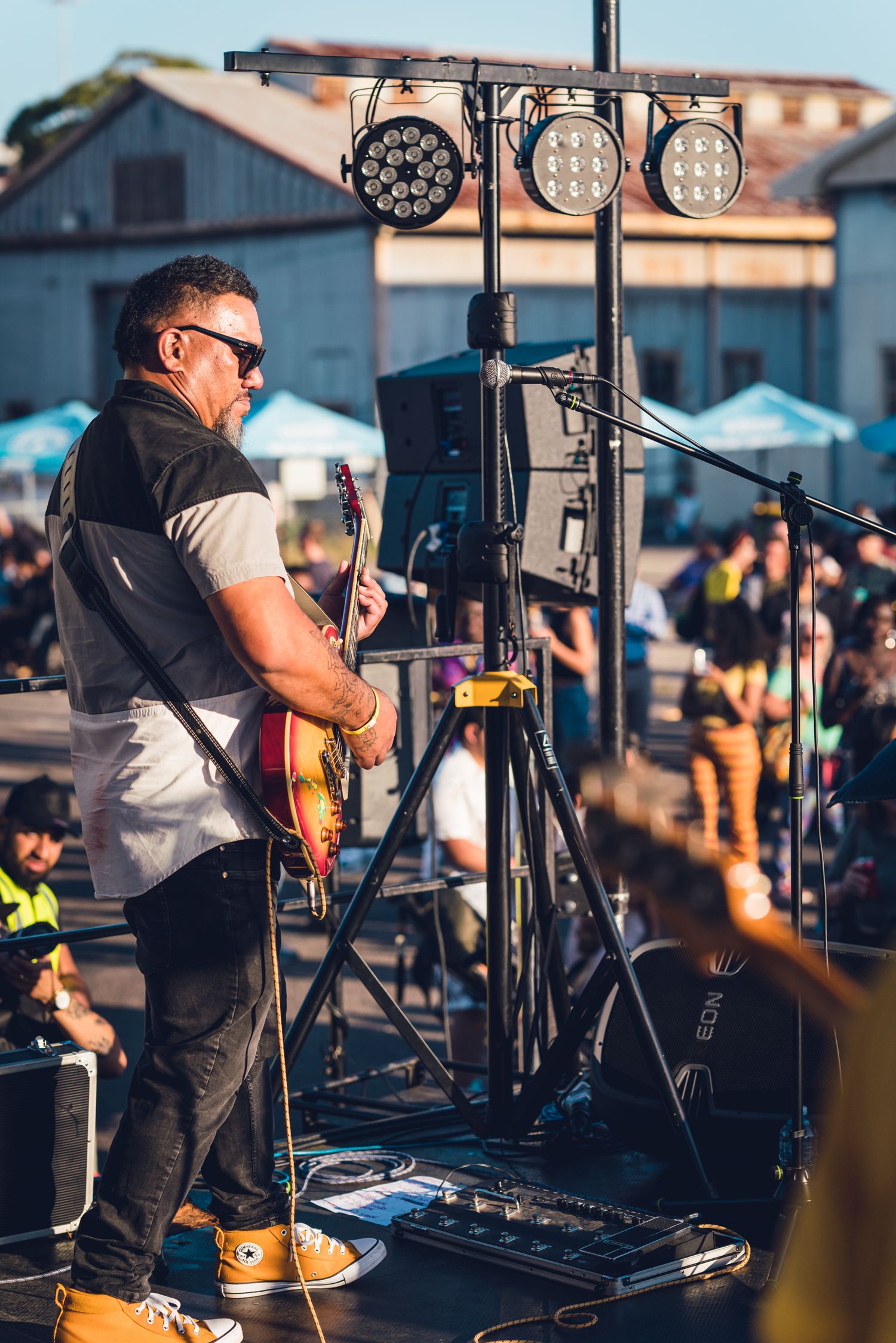 Guitarist in sunglasses playing electric guitar on outdoor festival stage with stage lighting.