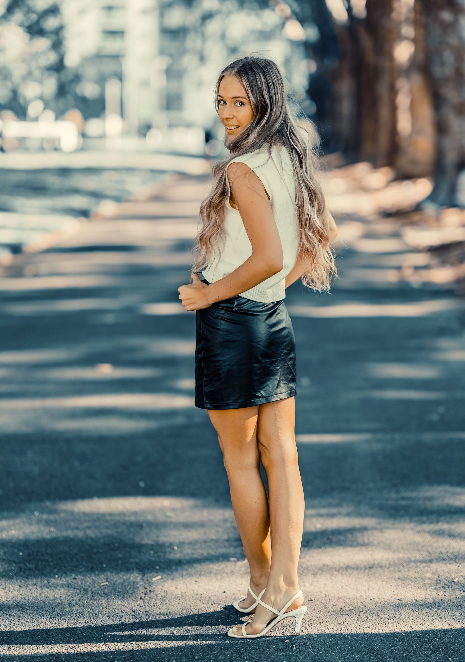 Young woman with long blonde hair wearing a black leather mini skirt and white heels posing outdoors.