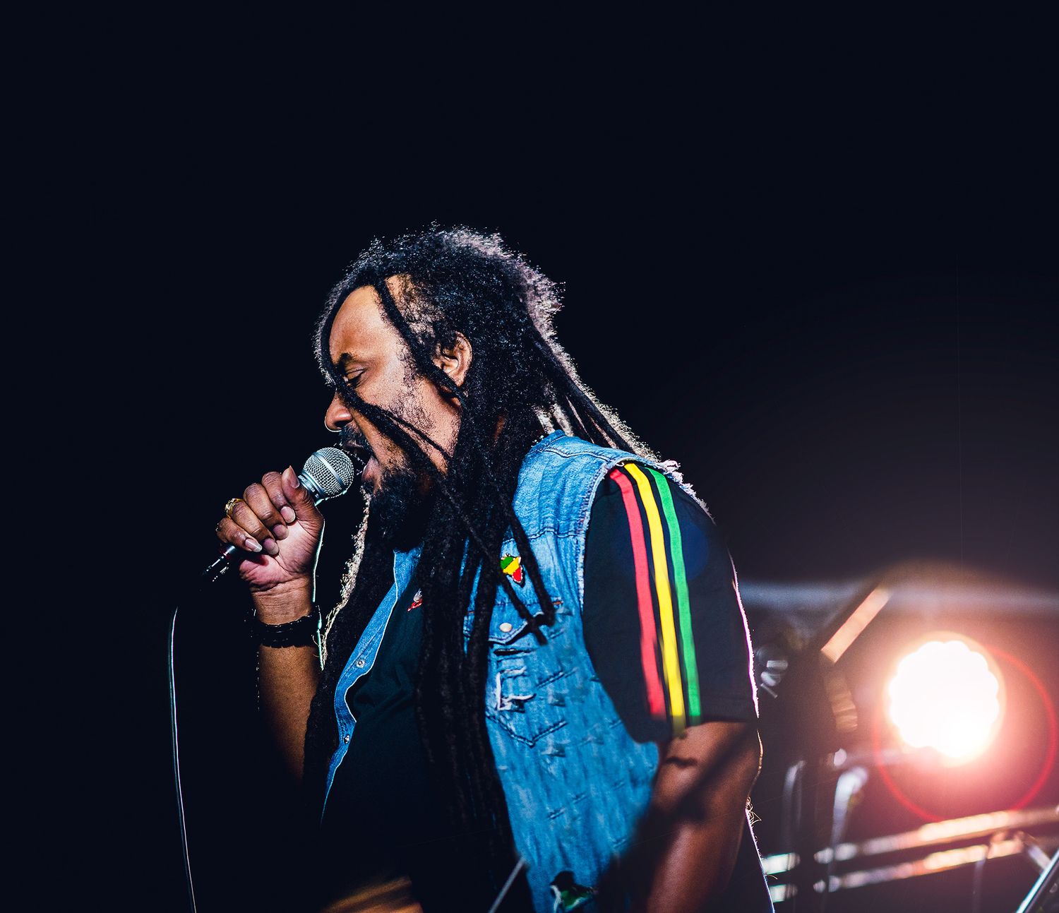 A reggae musician with dreadlocks performs on stage, singing into a microphone under dramatic lighting.