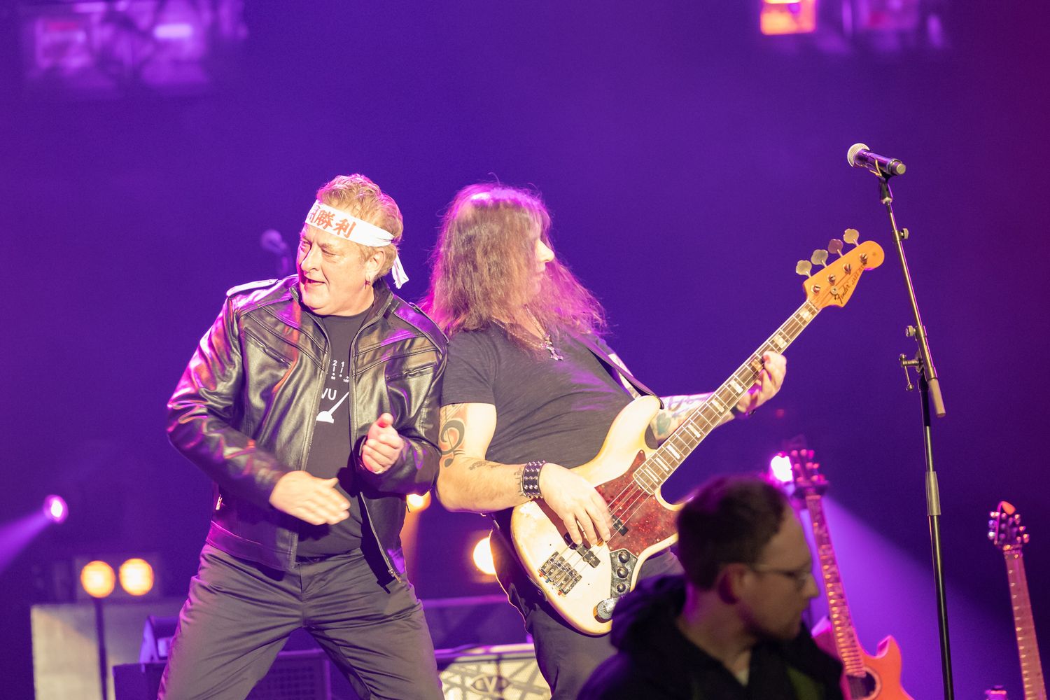 Two rock musicians perform energetically on a purple-lit stage, one singing and one playing bass guitar.