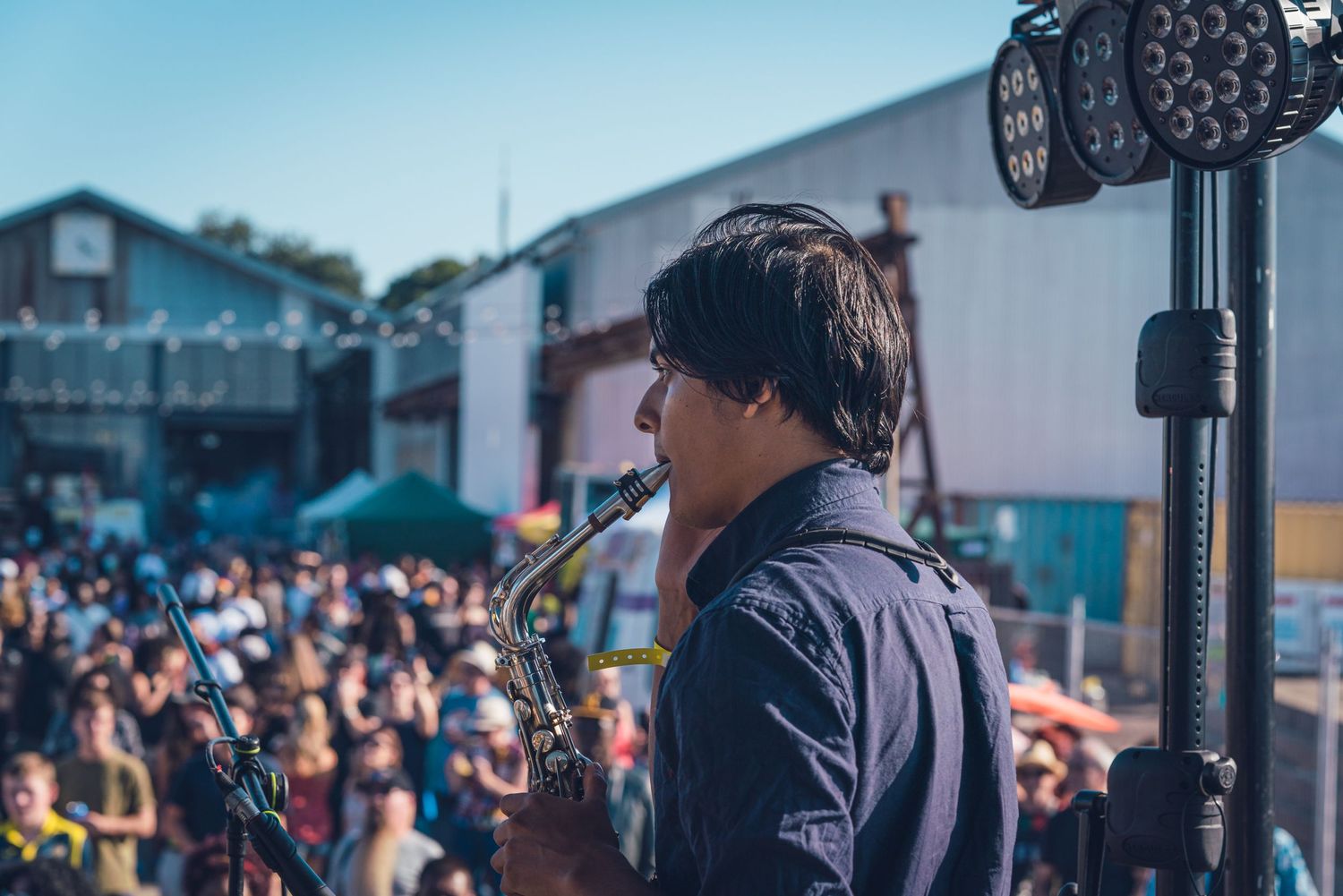 Saxophonist performing on outdoor festival stage with large crowd and blue sky in background.