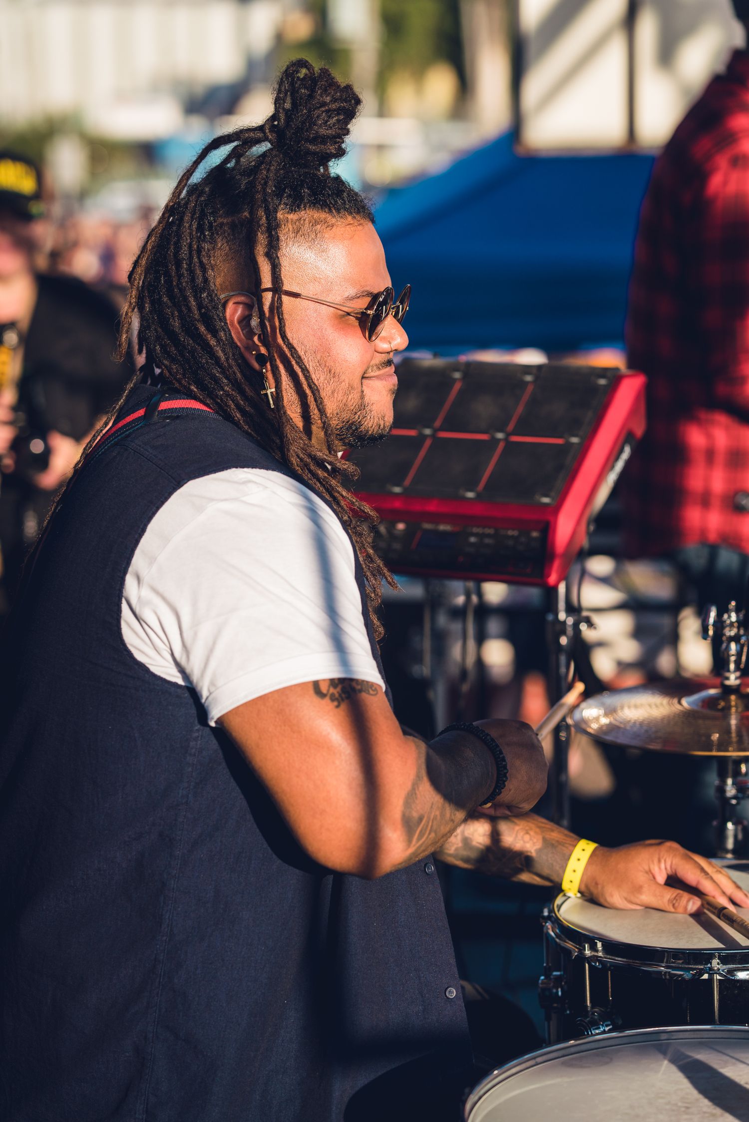 Drummer with dreadlocks and sunglasses playing snare drum at outdoor music festival.