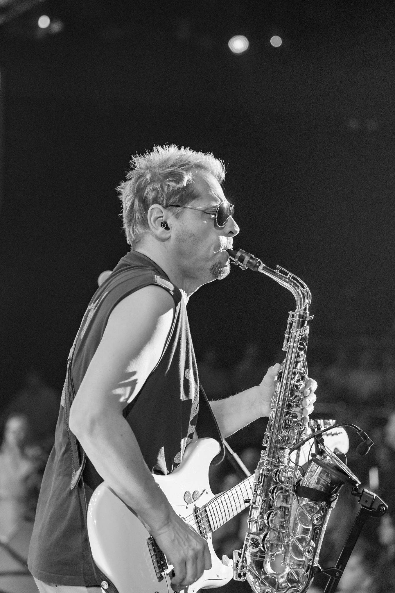 Black and white photo of a male saxophonist playing alto sax while holding an electric guitar on stage.