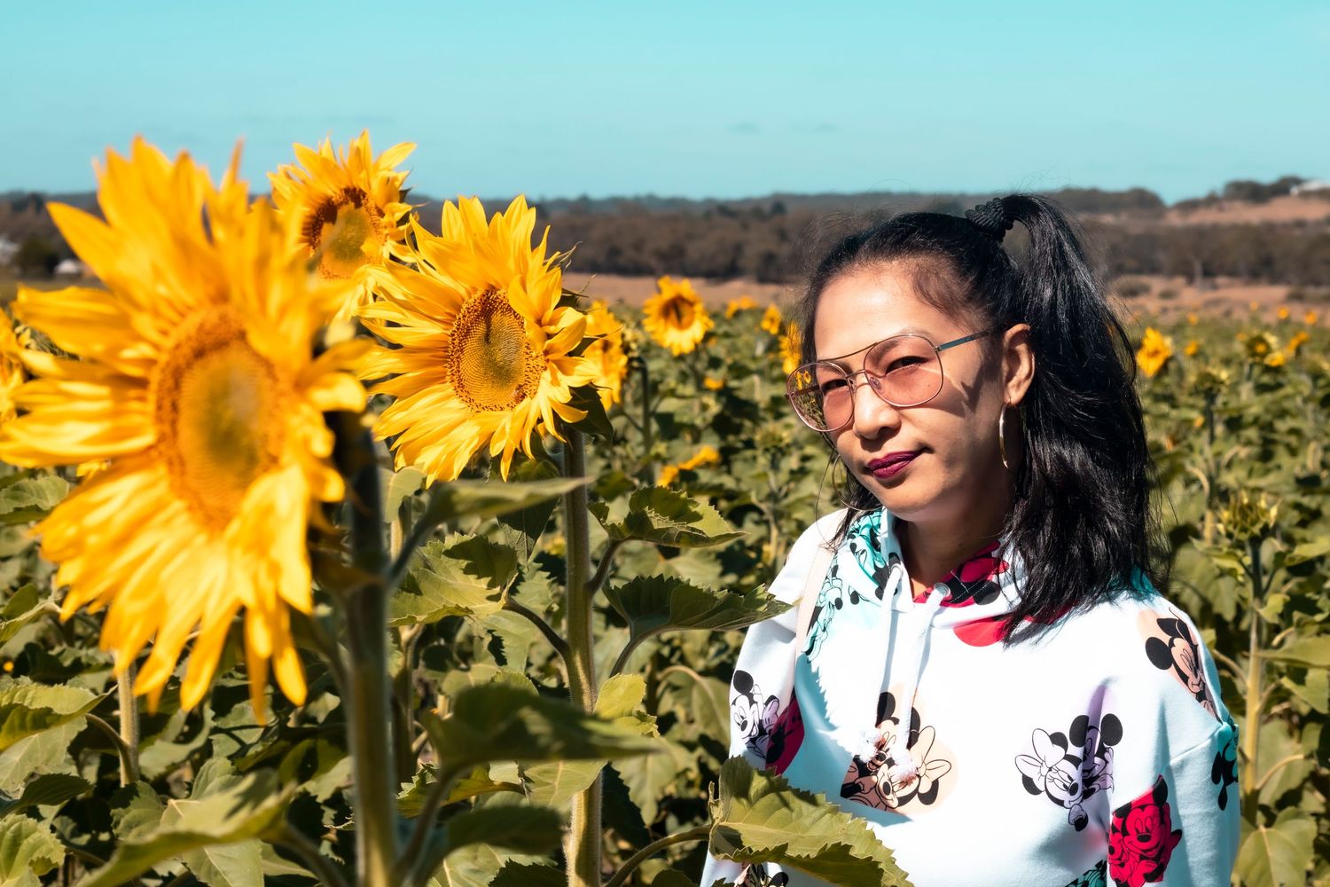 A young woman wearing glasses and a floral top stands in a sunny sunflower field outdoors.
