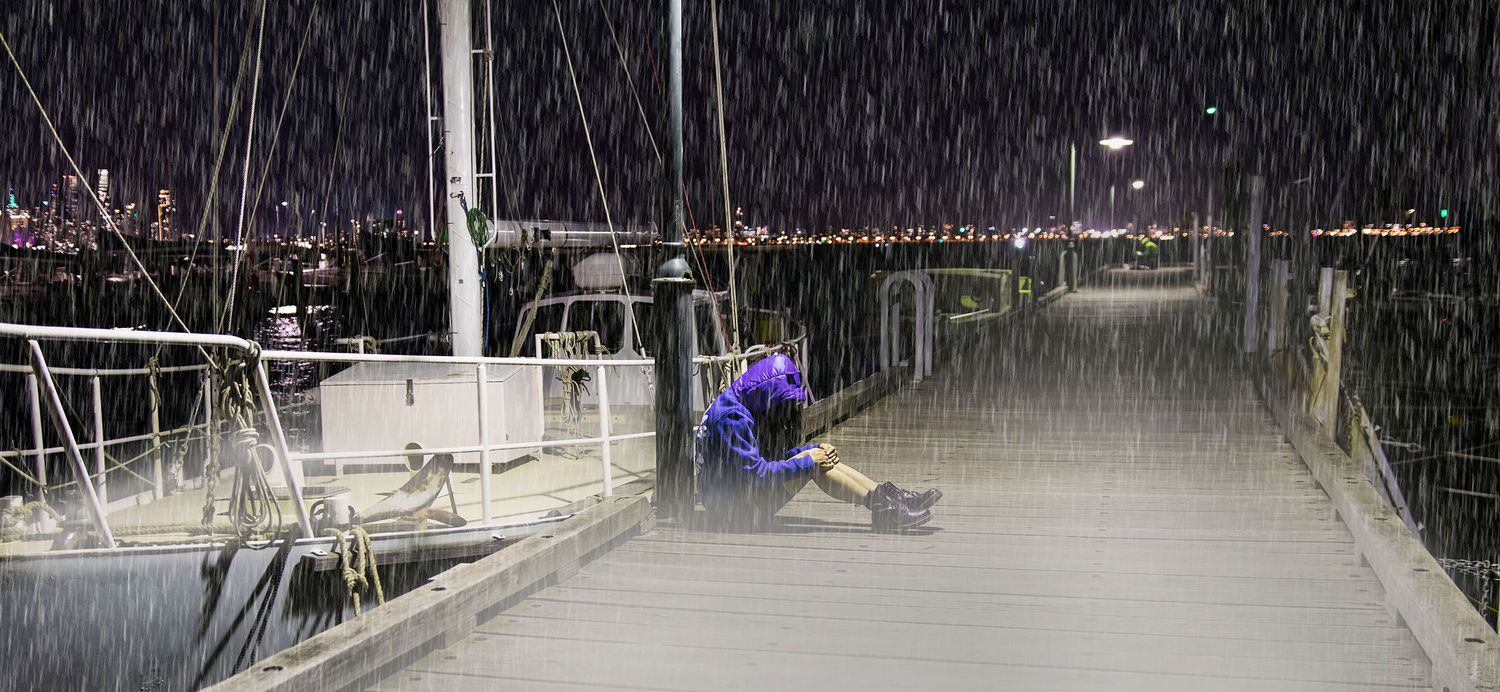 Person sitting alone on a wet dock during heavy rain at night with city lights reflecting on water.