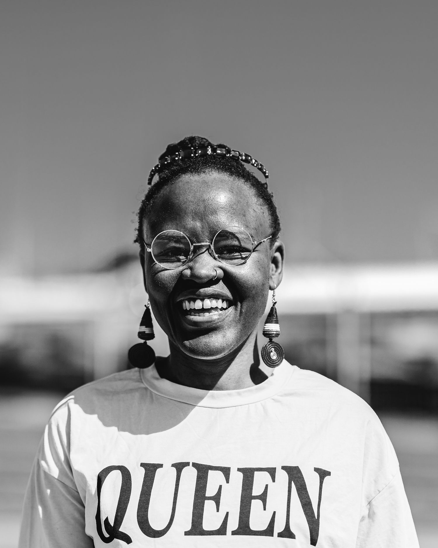Joyful woman in QUEEN shirt and round glasses smiling in black and white.