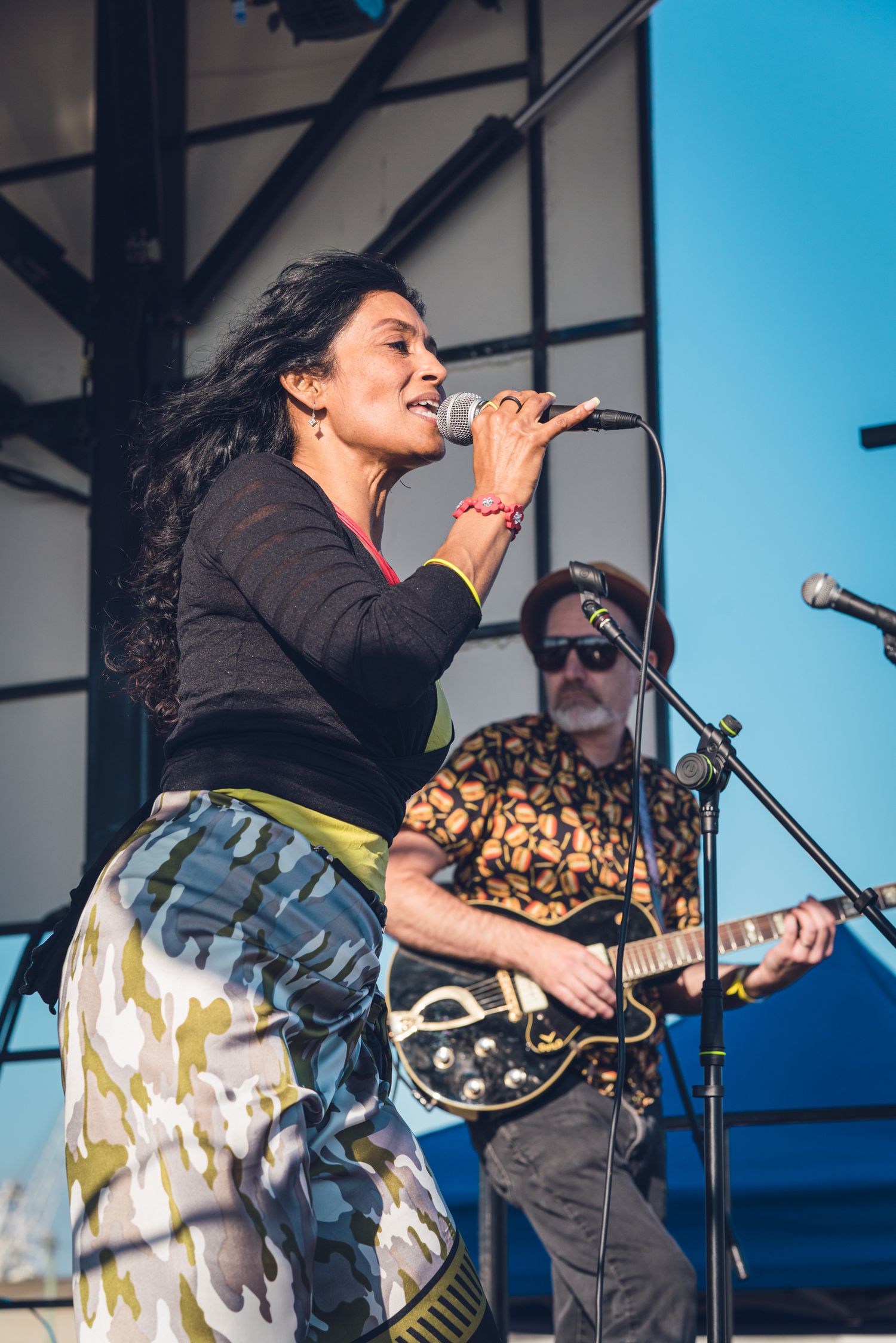 Female vocalist in camo skirt singing on outdoor stage with guitarist in floral shirt performing behind her.