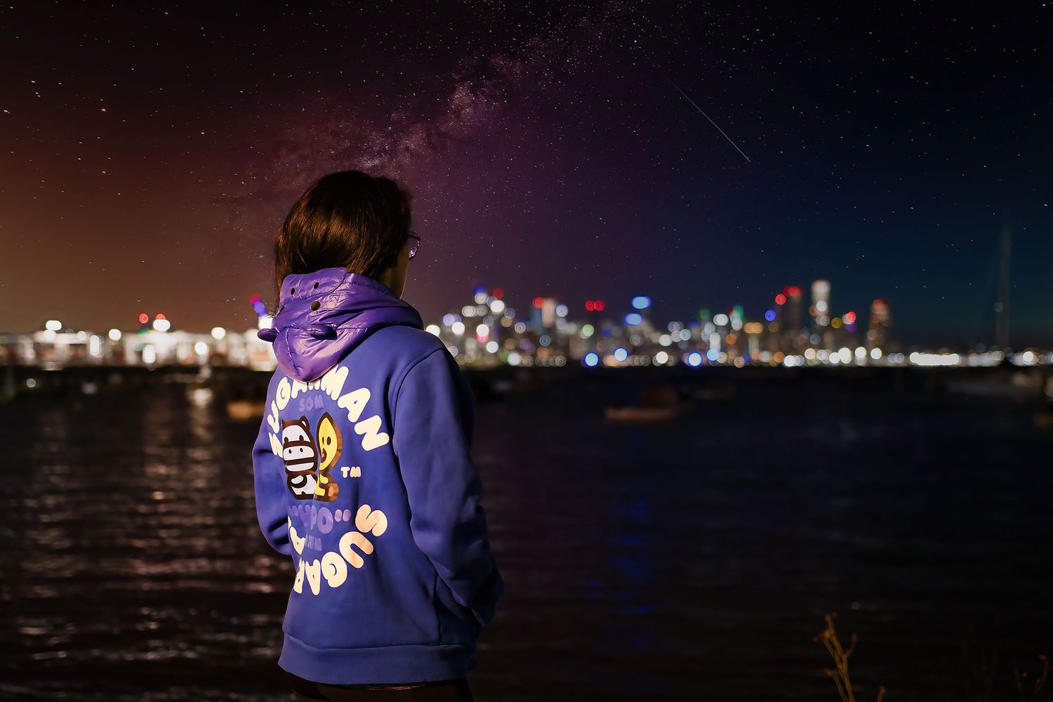 Person in purple hoodie gazes at illuminated city skyline reflected in water under a starry night sky.